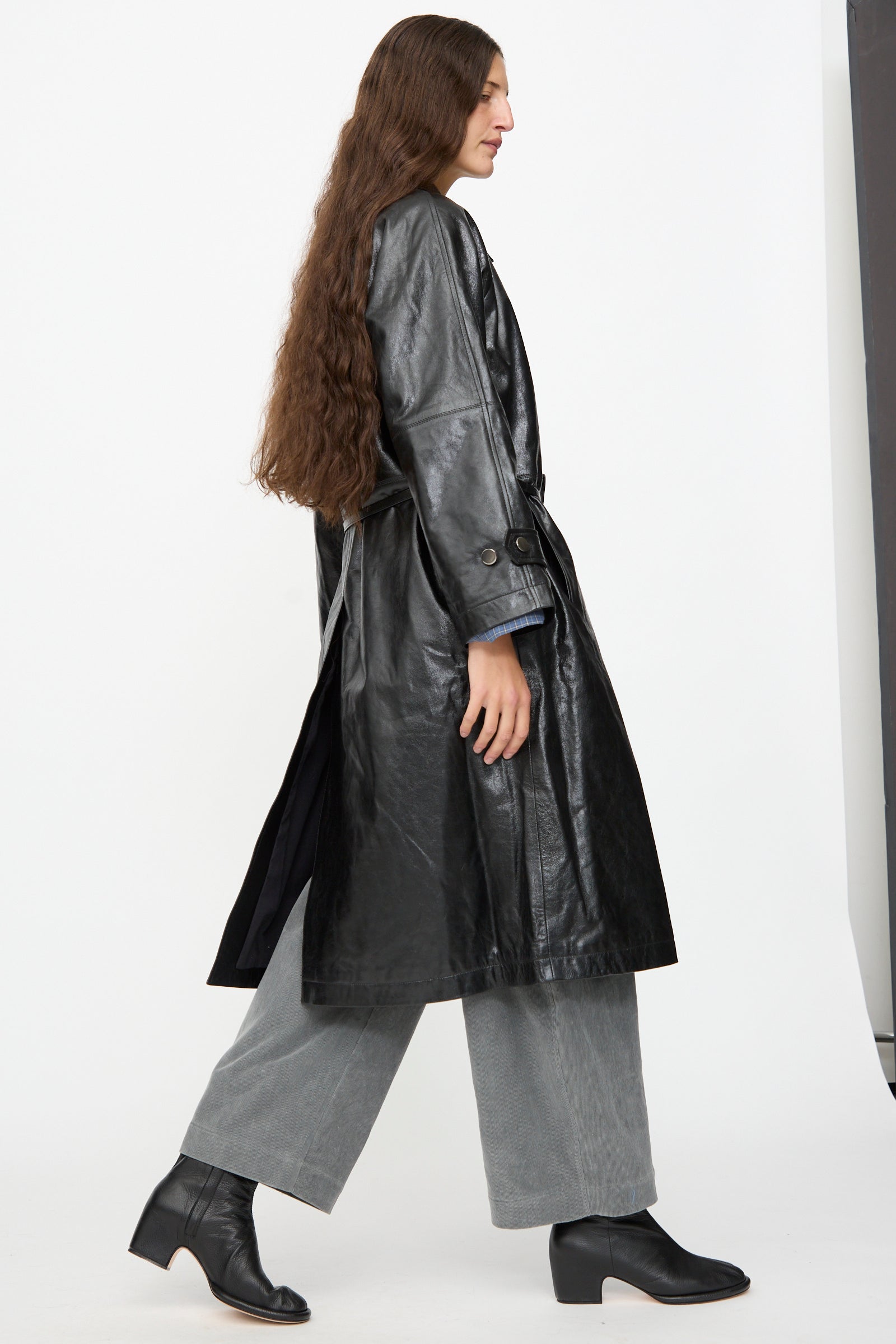 A person with long brown hair is wearing the Milena Silvano Leather Maria Trench in Black, paired with gray pants and black ankle boots, walking to the left against a plain white background.