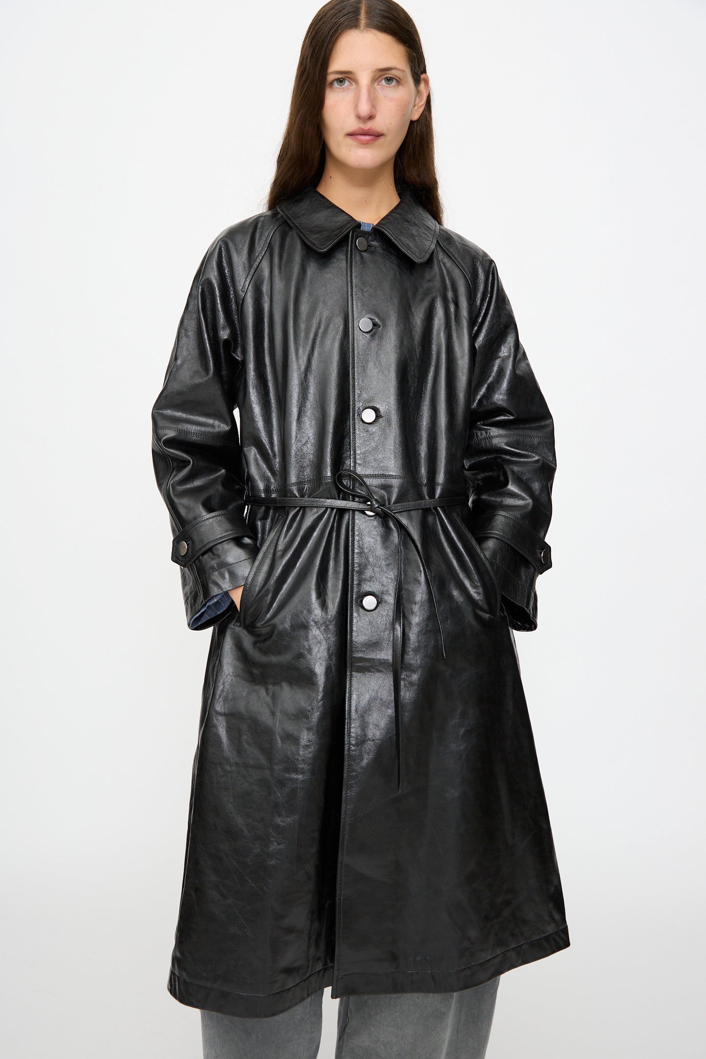 A person stands facing forward against a plain white background, wearing the Milena Silvano Leather Maria Trench in Black—crafted from vegetable tanned leather and featuring a belt—styled over grey pants.
