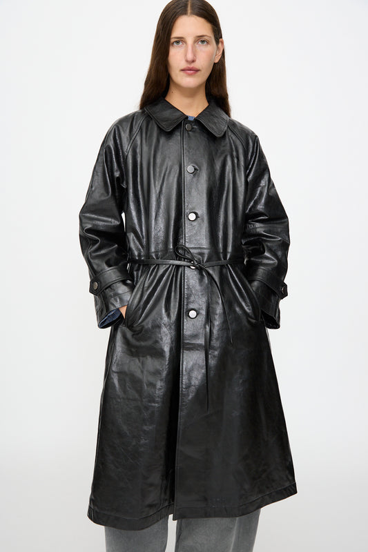 A person stands facing forward against a plain white background, wearing the Milena Silvano Leather Maria Trench in Black—crafted from vegetable tanned leather and featuring a belt—styled over grey pants.