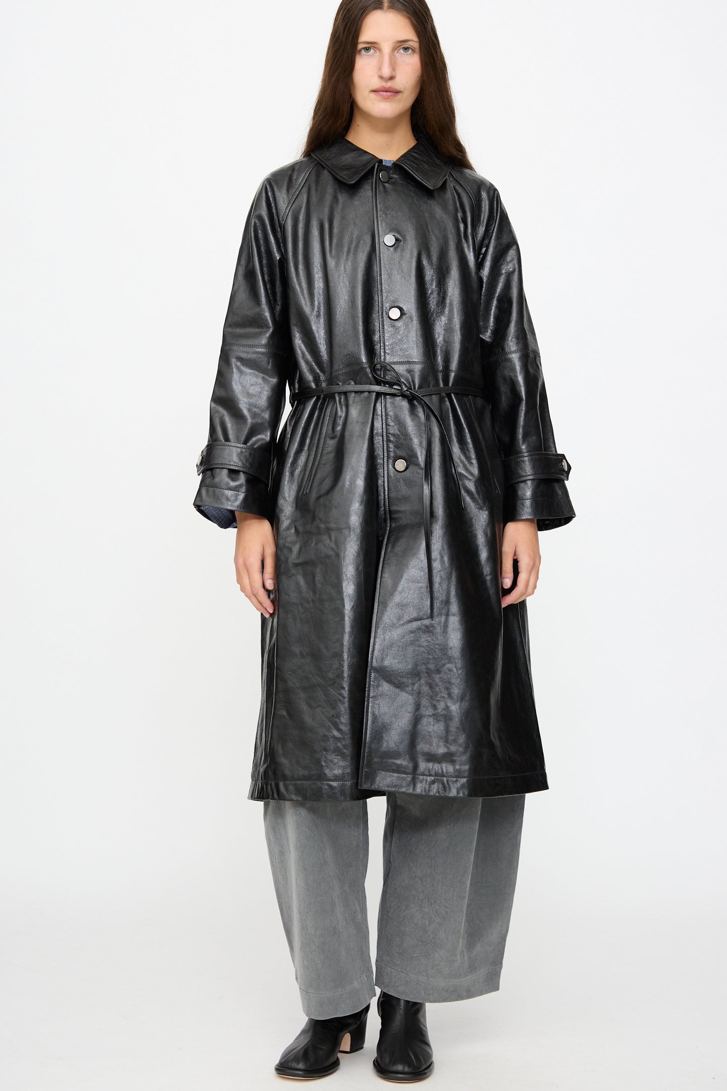 A person stands against a plain white background wearing the Milena Silvano Leather Maria Trench in Black, styled with wide gray pants and black shoes.