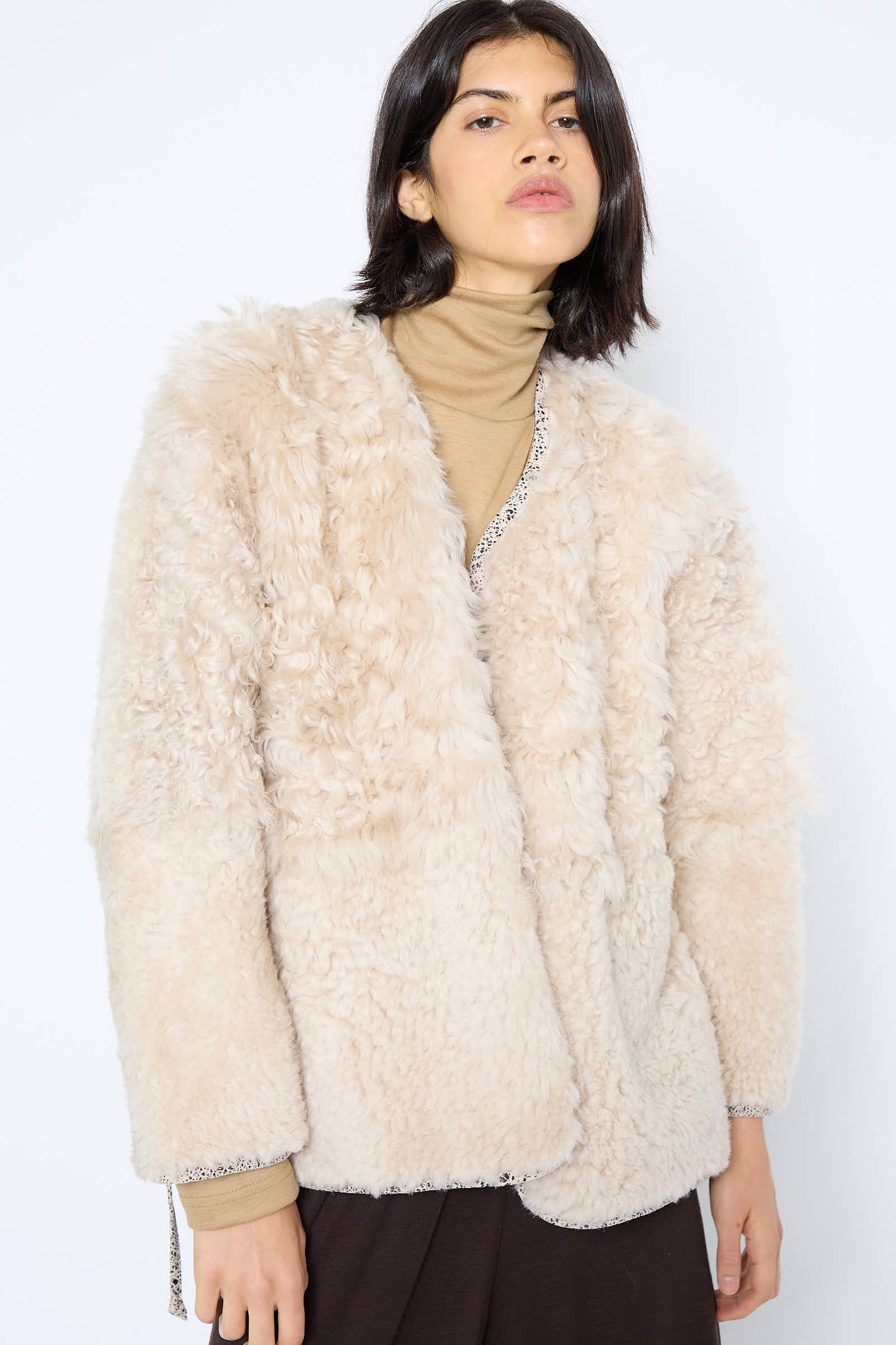 A person with medium-length dark hair wears a beige, fluffy Pyramid Robe in Classic by Milena Silvano over a tan turtleneck, against a plain white background.