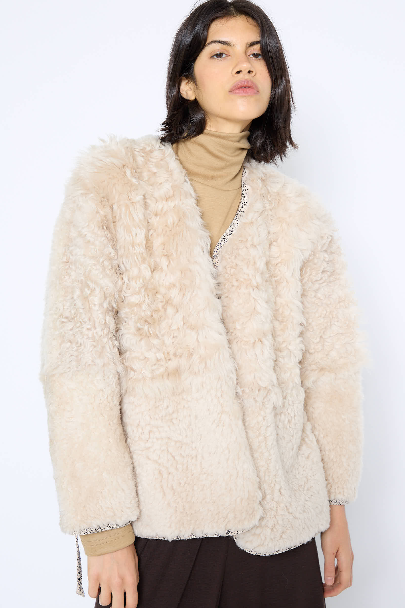 A person with medium-length dark hair wears a beige, fluffy Pyramid Robe in Classic by Milena Silvano over a tan turtleneck, against a plain white background.