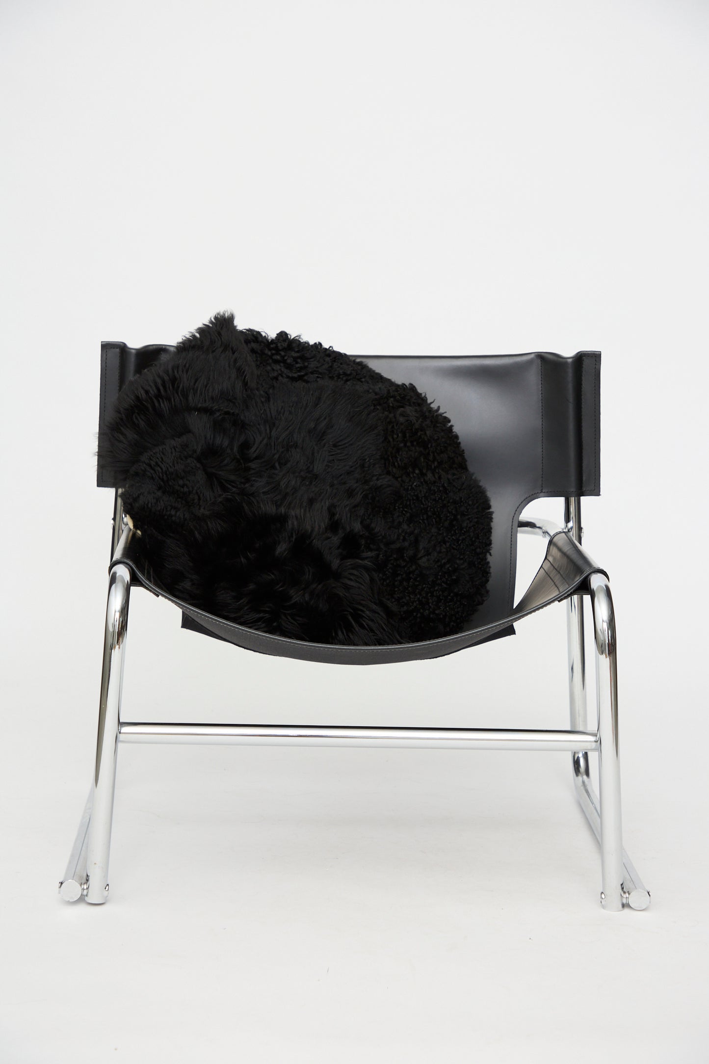 A modern black leather and chrome chair with a black fur throw draped over the seat is set against a plain white background, perfectly complemented by the Milena Silvano Shaggy Sheepskin Large Floor Pillow in Black for added comfort and style.