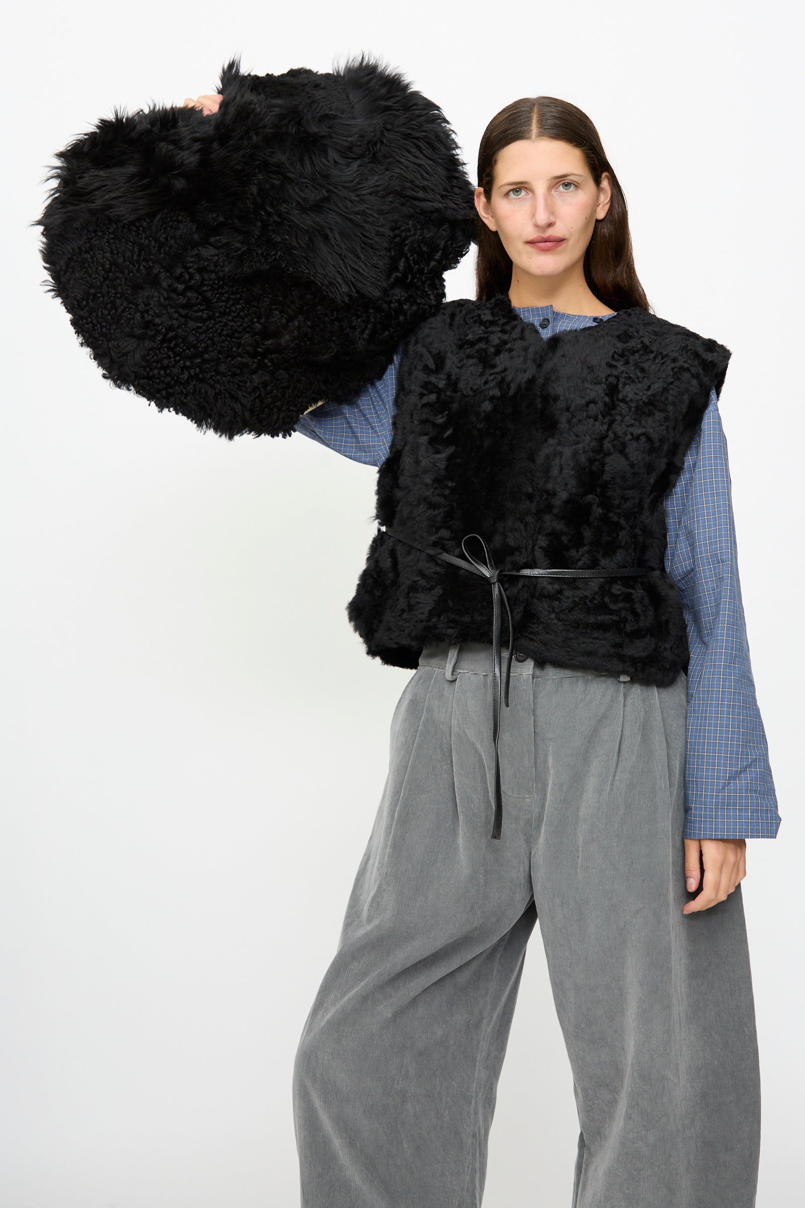 A woman wearing a black faux fur vest and gray wide-leg pants holds up the Milena Silvano Shaggy Sheepskin Large Floor Pillow in Black against a plain background.