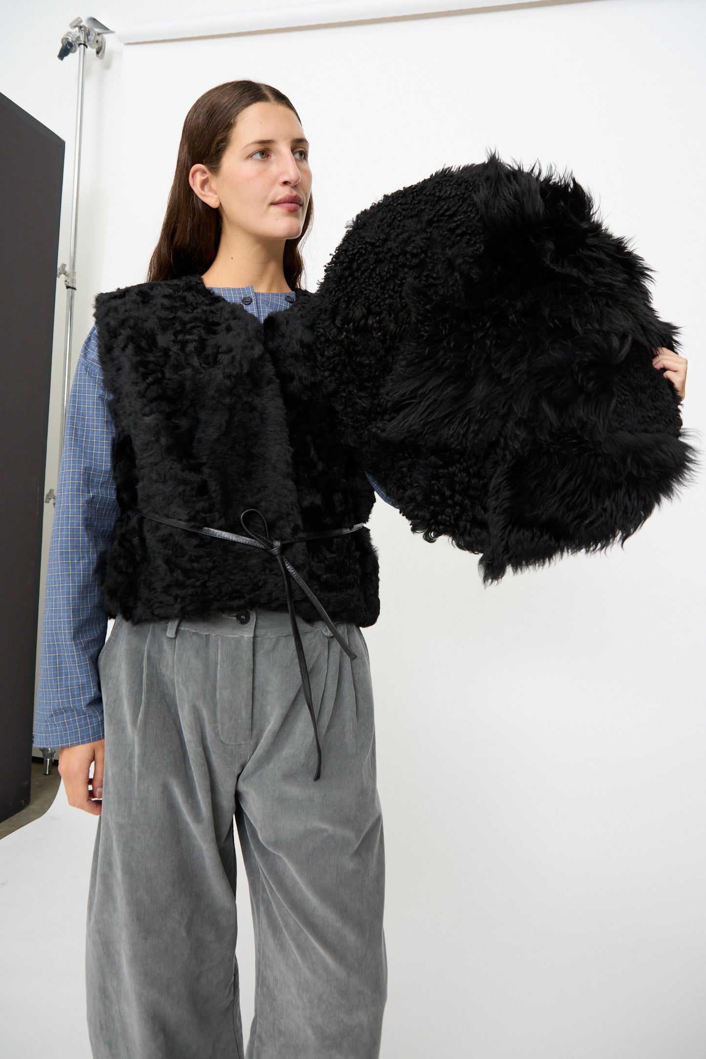 A person wearing a black faux fur vest and gray pants holds the Milena Silvano Shaggy Sheepskin Large Floor Pillow in Black against a plain white background.