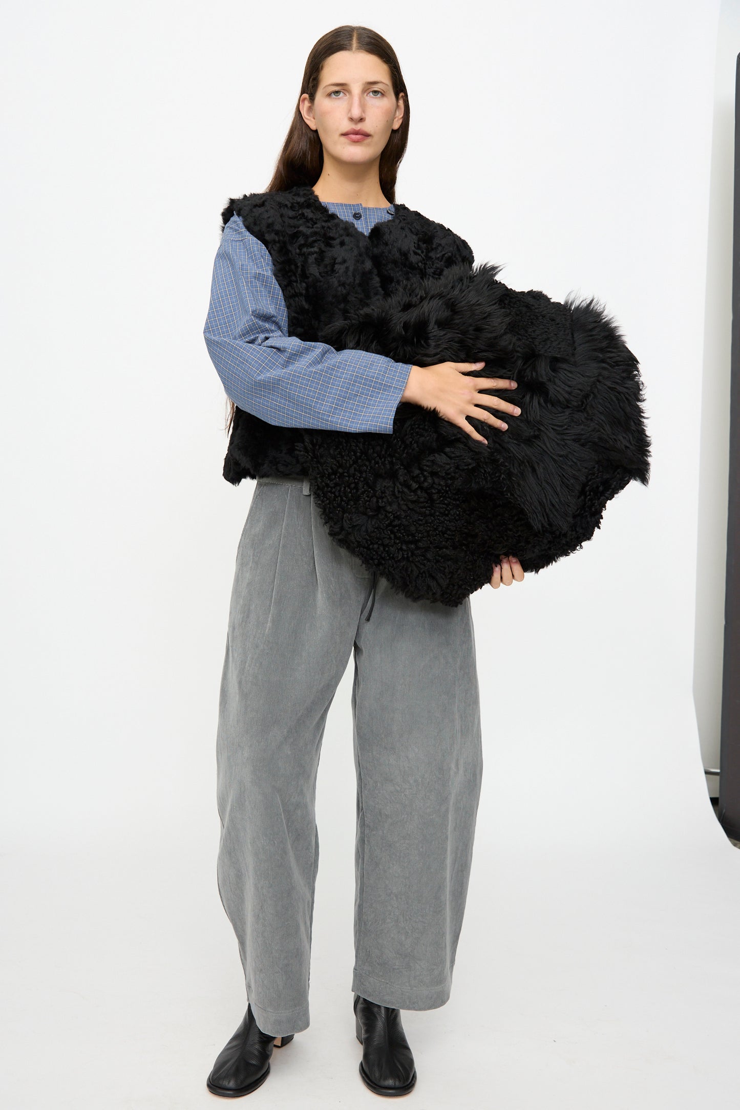 A person stands on a white background, wearing gray pants, a blue top, and black shoes, holding the Milena Silvano Shaggy Sheepskin Large Floor Pillow in Black.