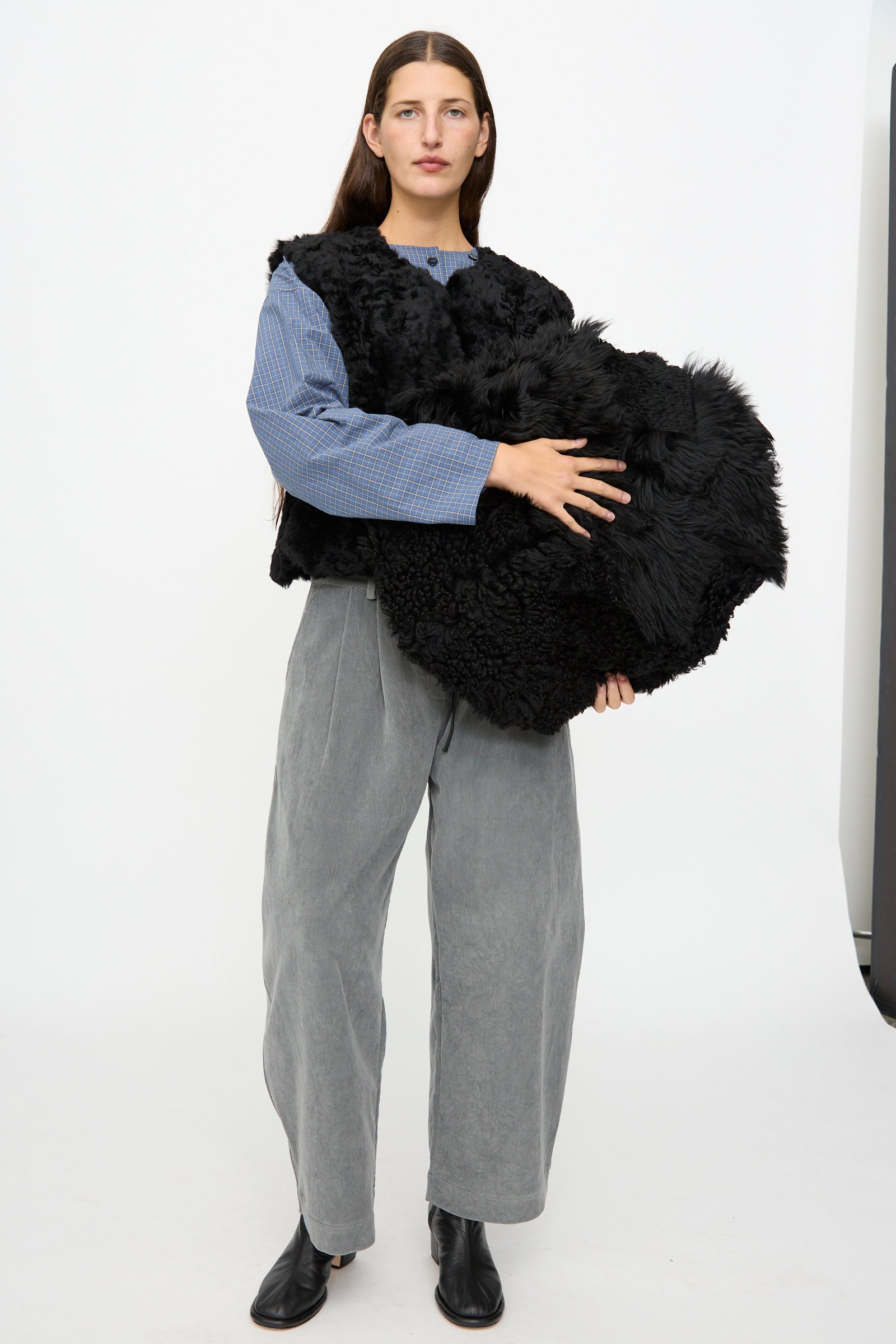 A person stands on a white background, wearing gray pants, a blue top, and black shoes, holding the Milena Silvano Shaggy Sheepskin Large Floor Pillow in Black.