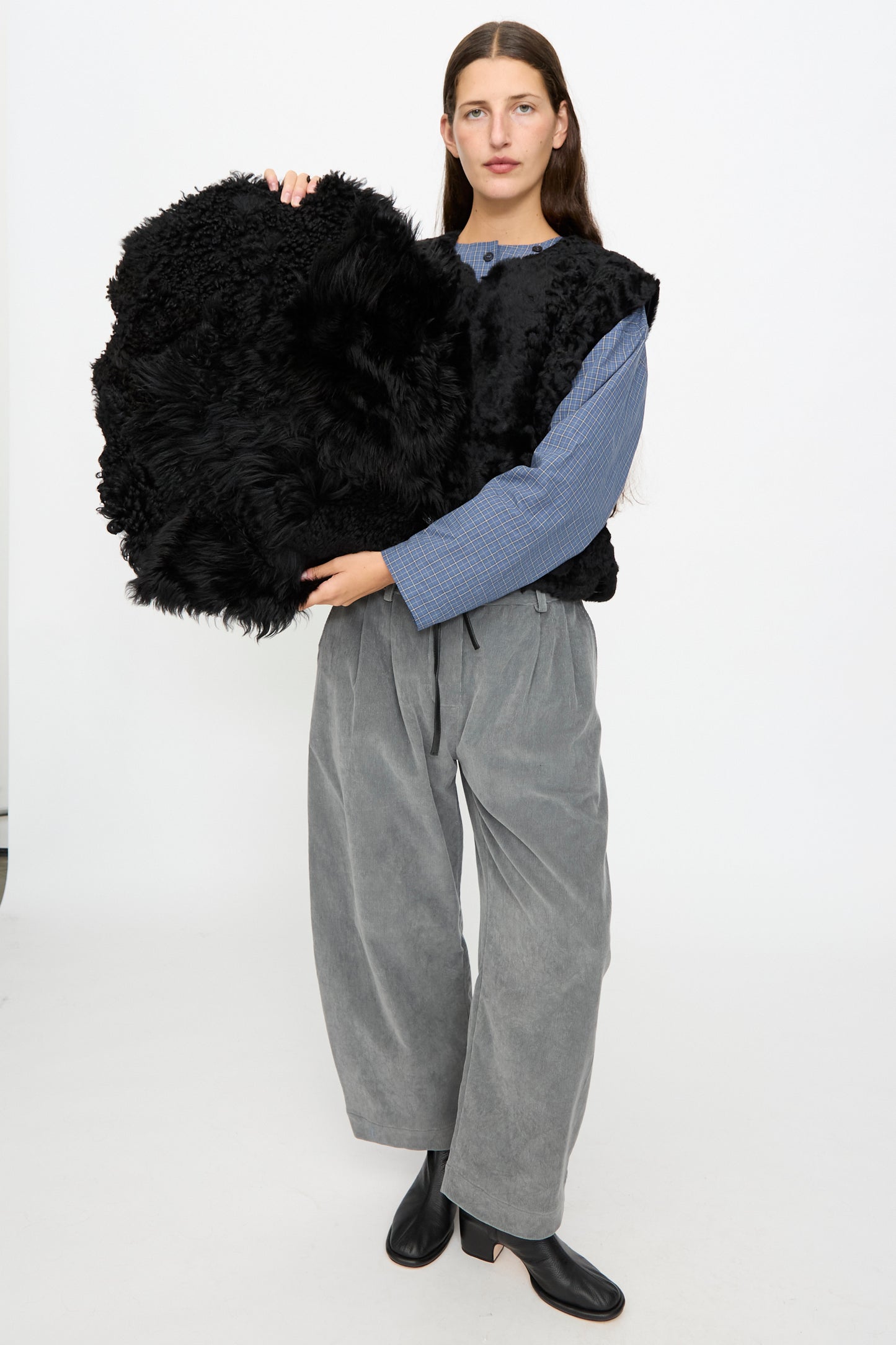 A person in loose gray pants, black shoes, and a fuzzy black vest holds the Milena Silvano Shaggy Sheepskin Large Floor Pillow in Black against a plain white background.