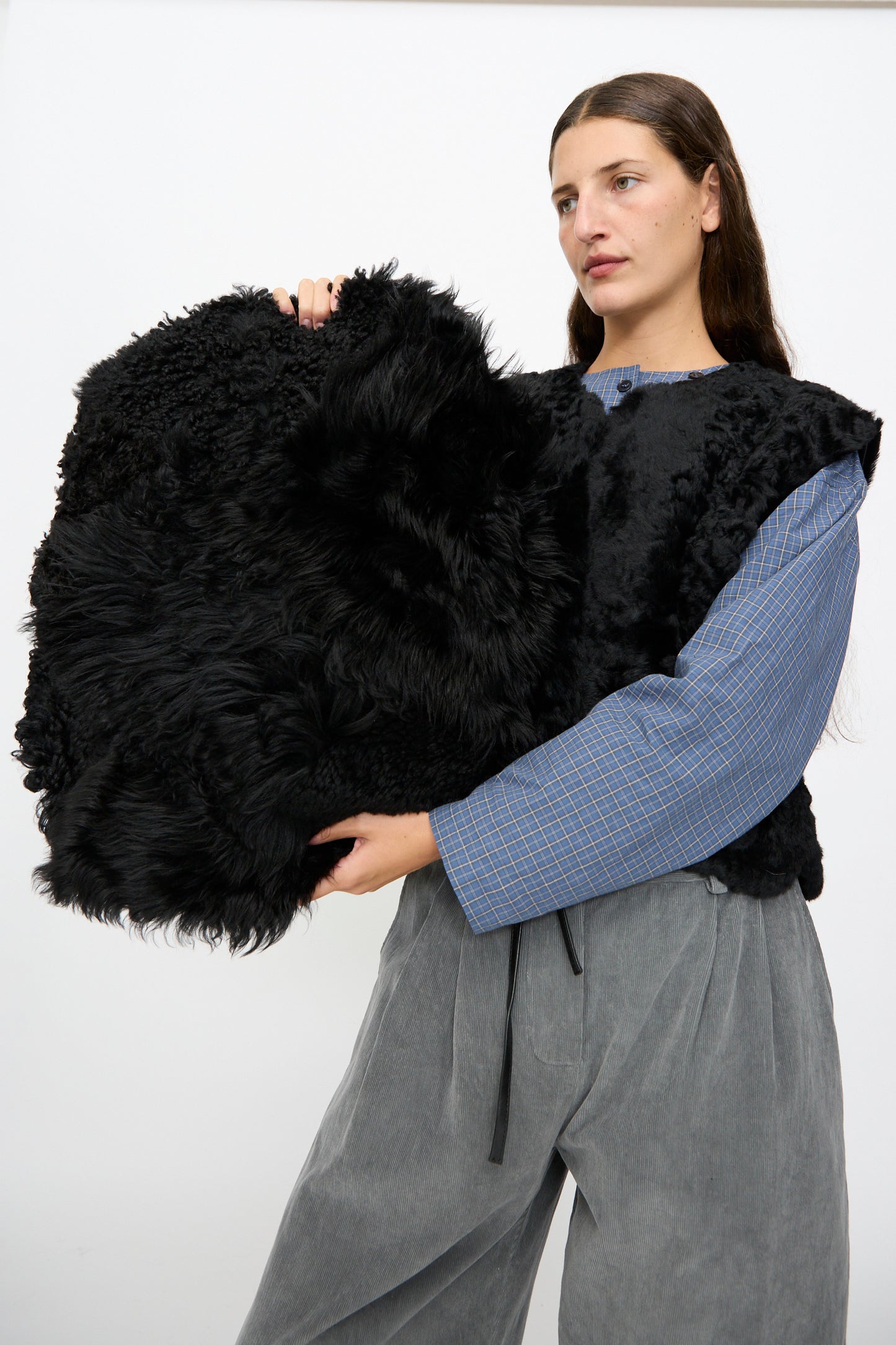 A person in gray pants and a blue plaid top holds the Milena Silvano Shaggy Sheepskin Large Floor Pillow in Black against a plain white background. This unique, limited edition piece infuses any space with its luxurious texture.