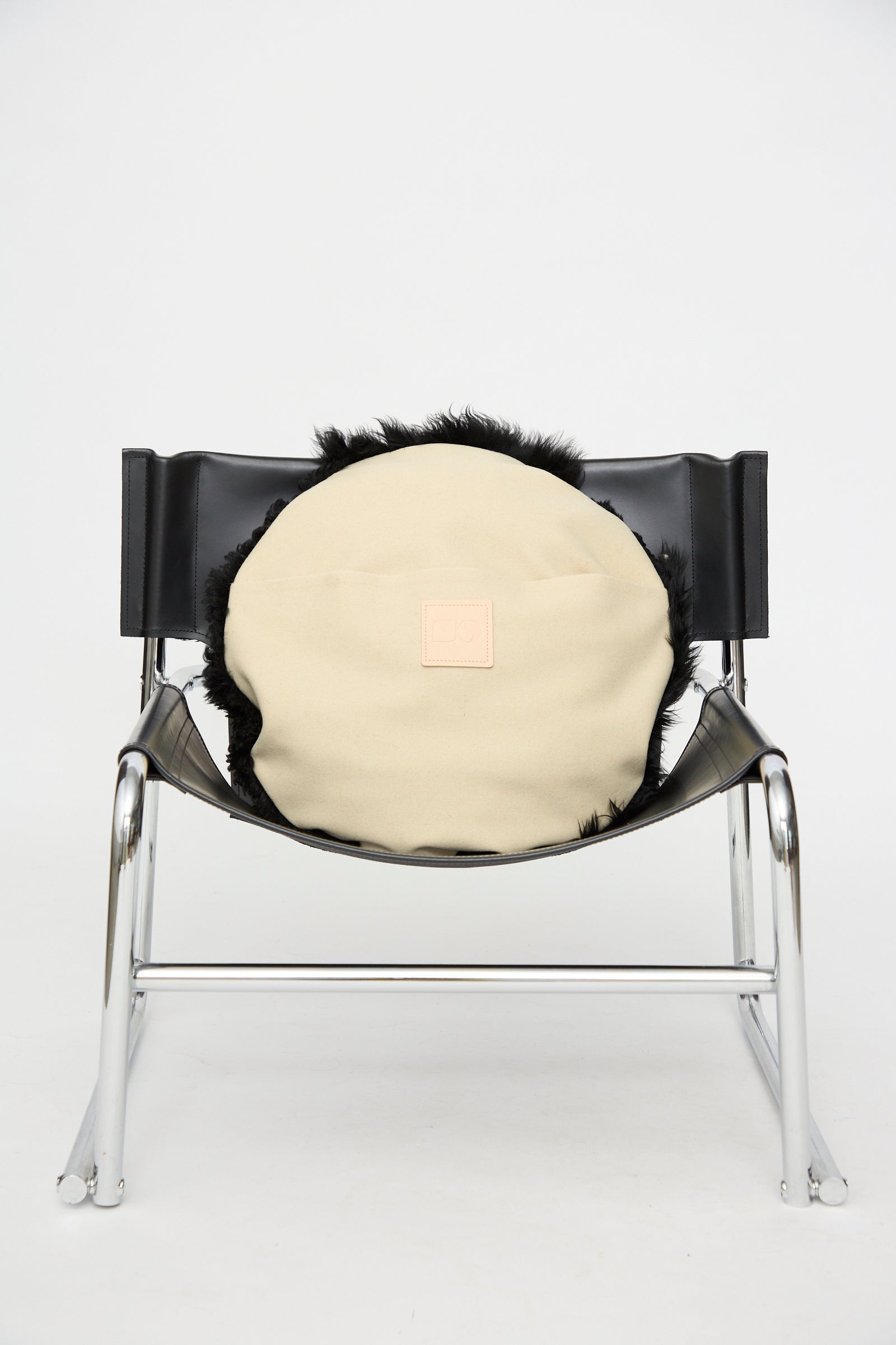 A black leather and chrome chair features the Shaggy Sheepskin Large Floor Pillow in Black by Milena Silvano, placed on the seat and photographed against a plain white background.