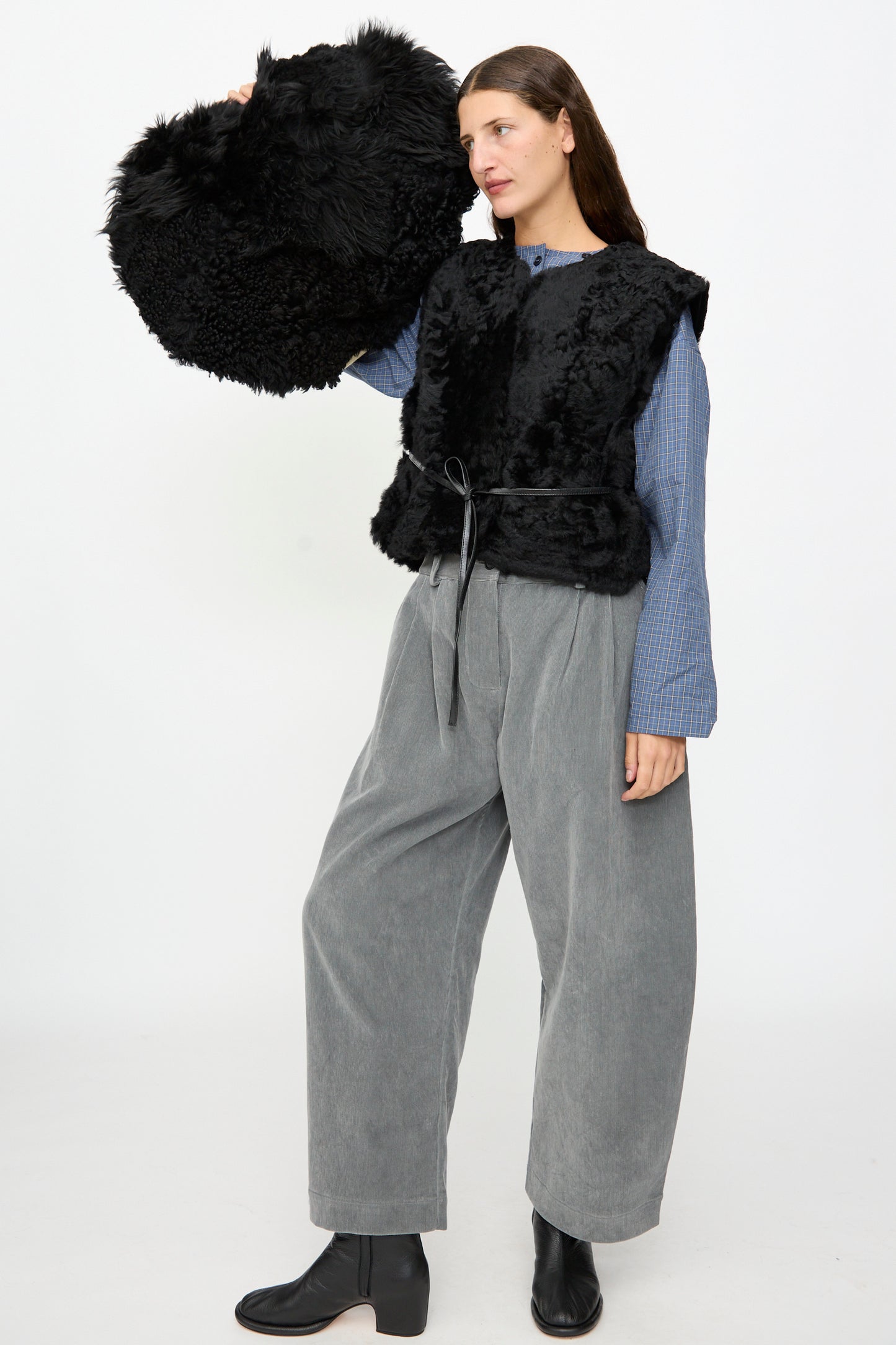 A woman wearing wide grey pants, a blue shirt, and a black fur vest holds the Milena Silvano Shaggy Sheepskin Large Floor Pillow in Black against a plain white background.