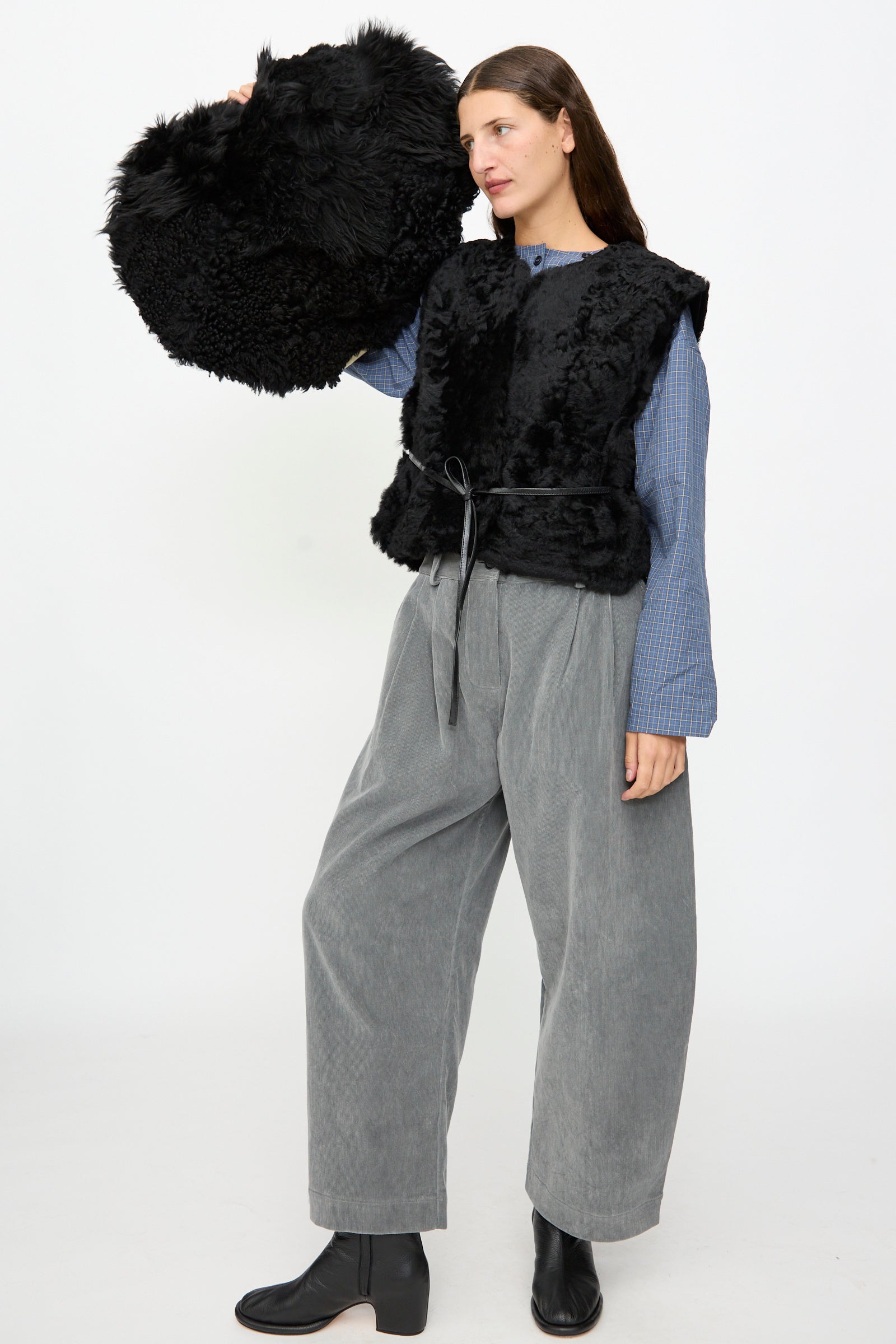 A woman wearing wide grey pants, a blue shirt, and a black fur vest holds the Milena Silvano Shaggy Sheepskin Large Floor Pillow in Black against a plain white background.