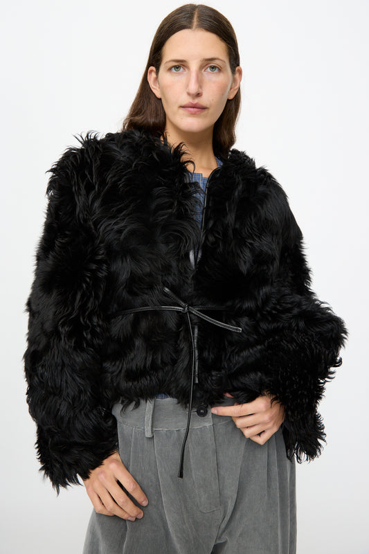 A person wears the Milena Silvano Shaggy Sheepskin Rainer Jacket in Black with gray corduroy pants, standing against a plain white background.