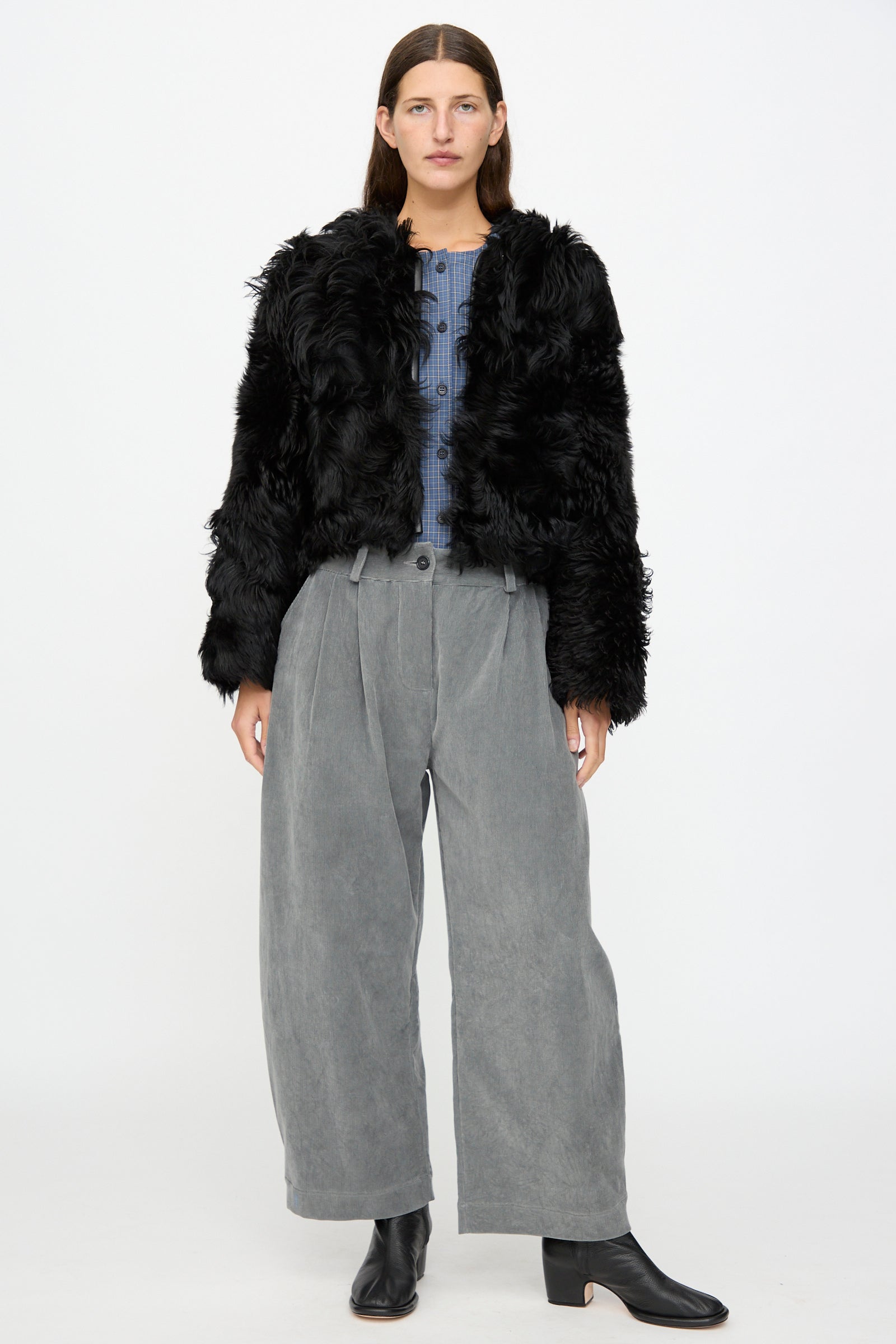 A person wears the Milena Silvano Shaggy Sheepskin Rainer Jacket in Black over a blue button-up shirt, paired with loose gray wide-leg pants and black ankle boots, standing against a plain white background.