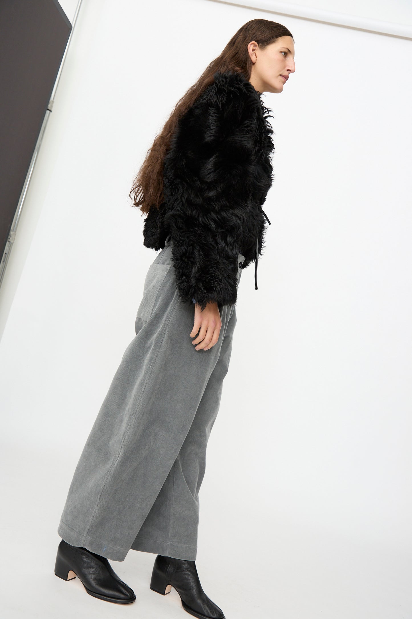 A person stands sideways against a plain white background, wearing the Milena Silvano Shaggy Sheepskin Rainer Jacket in Black, wide-leg gray pants, and black ankle boots.