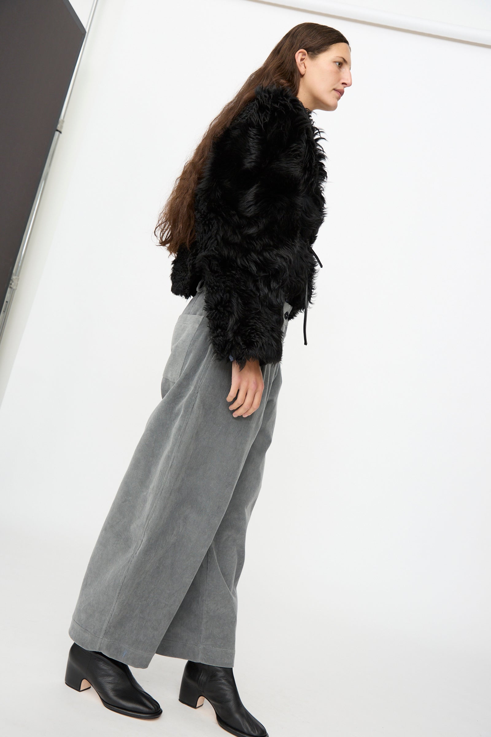 A person stands sideways against a plain white background, wearing the Milena Silvano Shaggy Sheepskin Rainer Jacket in Black, wide-leg gray pants, and black ankle boots.