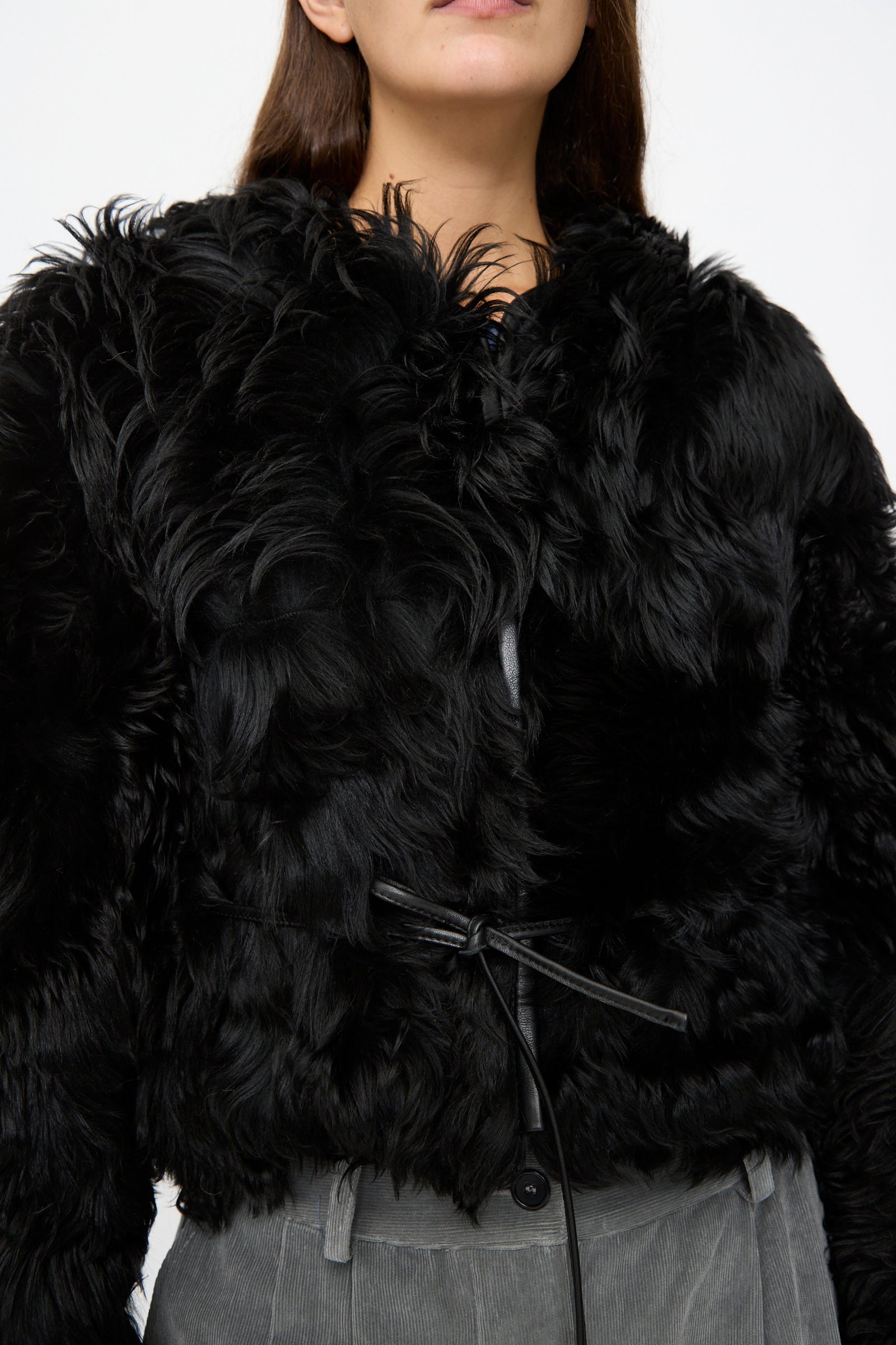 A person wearing the Milena Silvano Shaggy Sheepskin Rainer Jacket in Black, cinched with a thin belt and styled with gray pants. The photo is cropped from the shoulders to the hips.