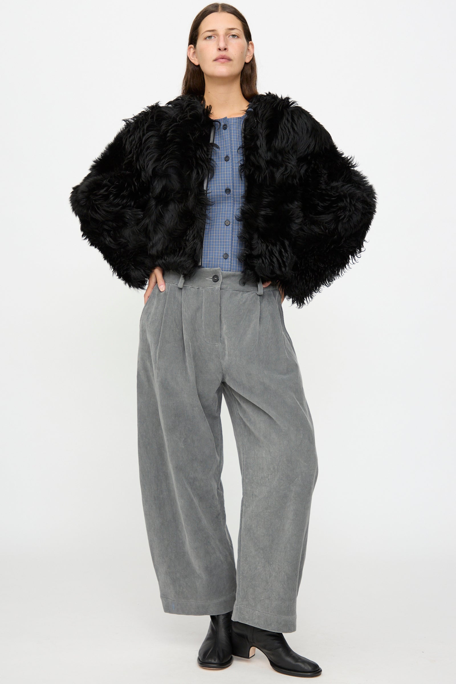 A person stands against a plain white background wearing the Milena Silvano Shaggy Sheepskin Rainer Jacket in Black, paired with a blue buttoned shirt, wide gray trousers, and black ankle boots.