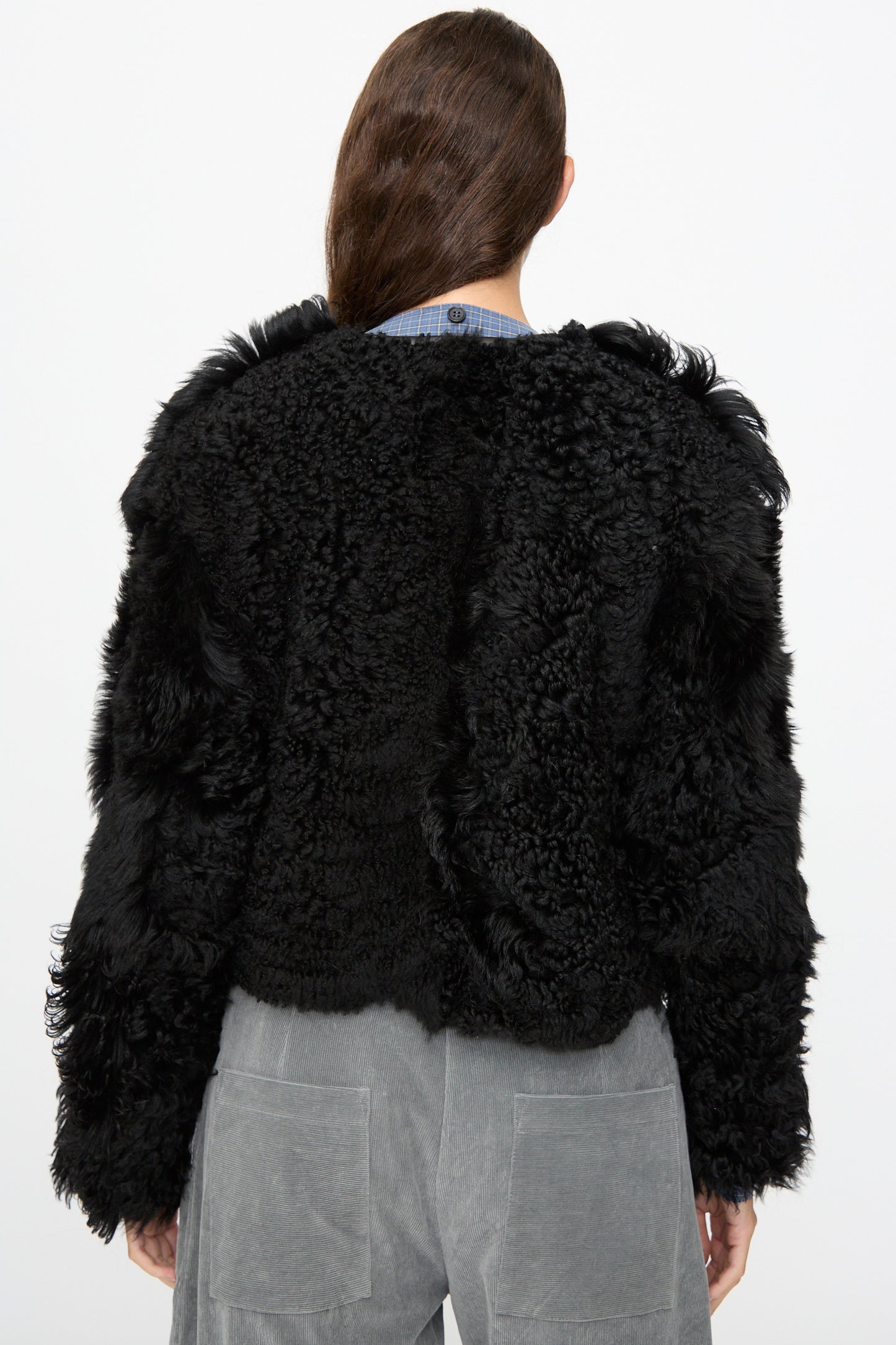 A person with long brown hair is shown from the back against a plain white background, wearing the Milena Silvano Shaggy Sheepskin Rainer Jacket in Black and gray corduroy pants.