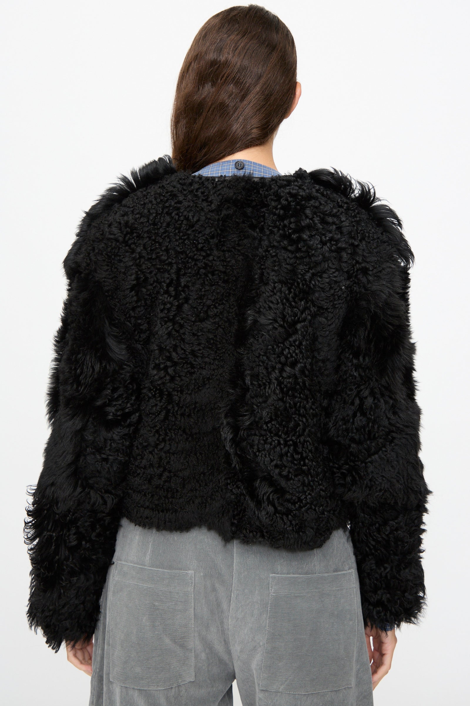 A person with long brown hair is shown from the back against a plain white background, wearing the Milena Silvano Shaggy Sheepskin Rainer Jacket in Black and gray corduroy pants.