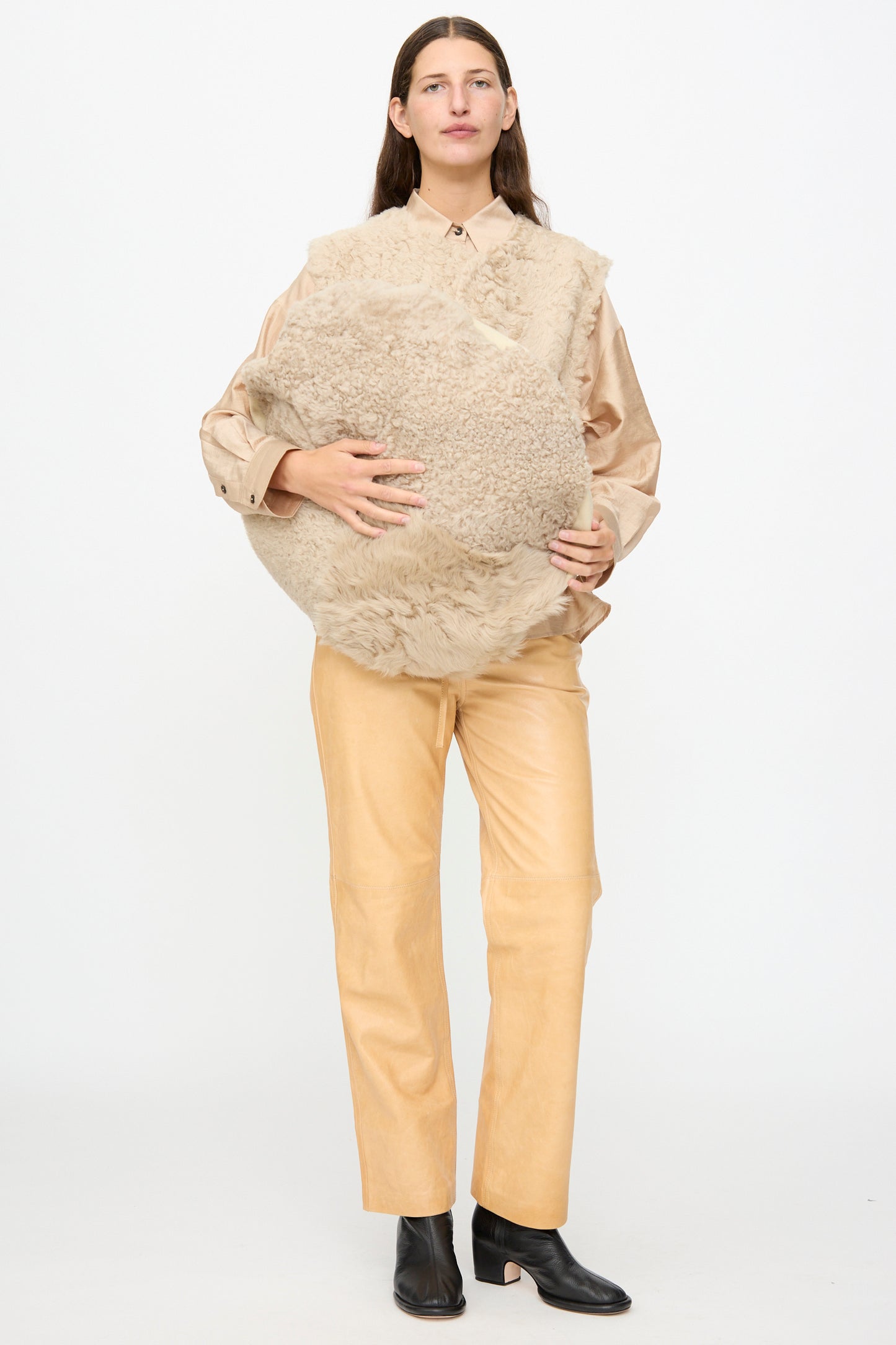 A person stands against a white background, wearing a beige shirt and pants with black boots, holding the Milena Silvano Sheepskin Large Floor Pillow in Oat.