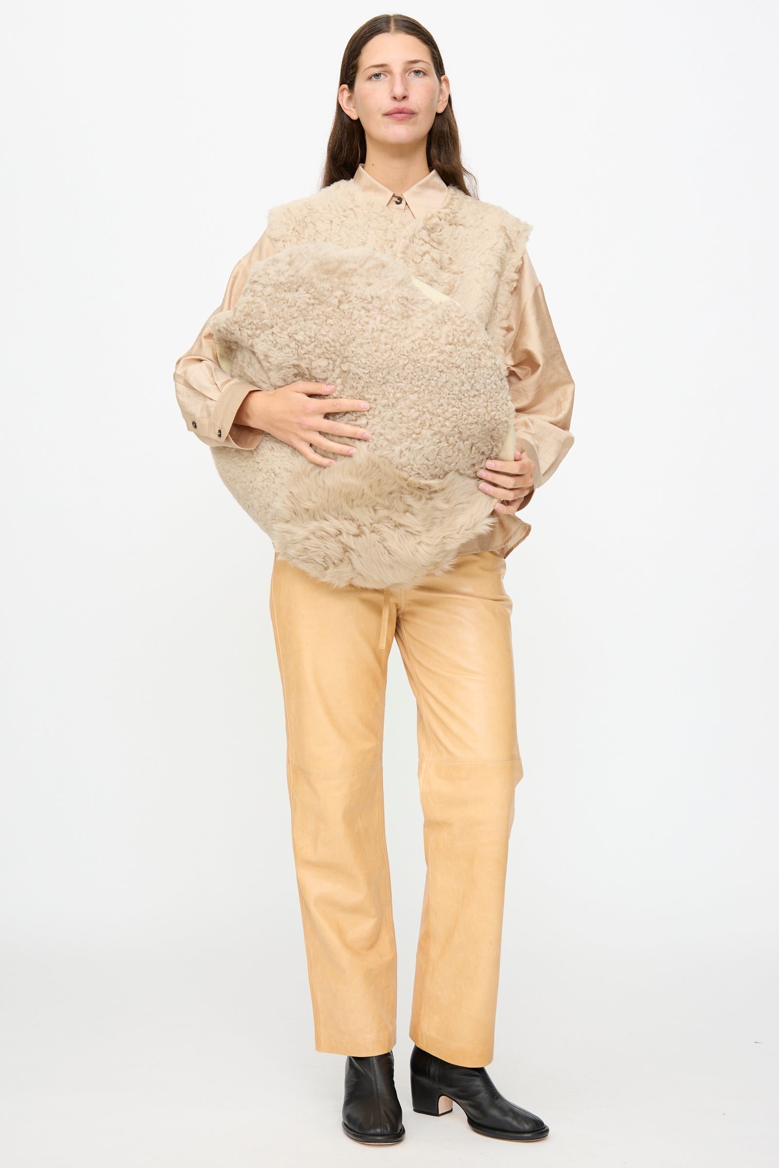 A person stands against a white background, wearing a beige shirt and pants with black boots, holding the Milena Silvano Sheepskin Large Floor Pillow in Oat.