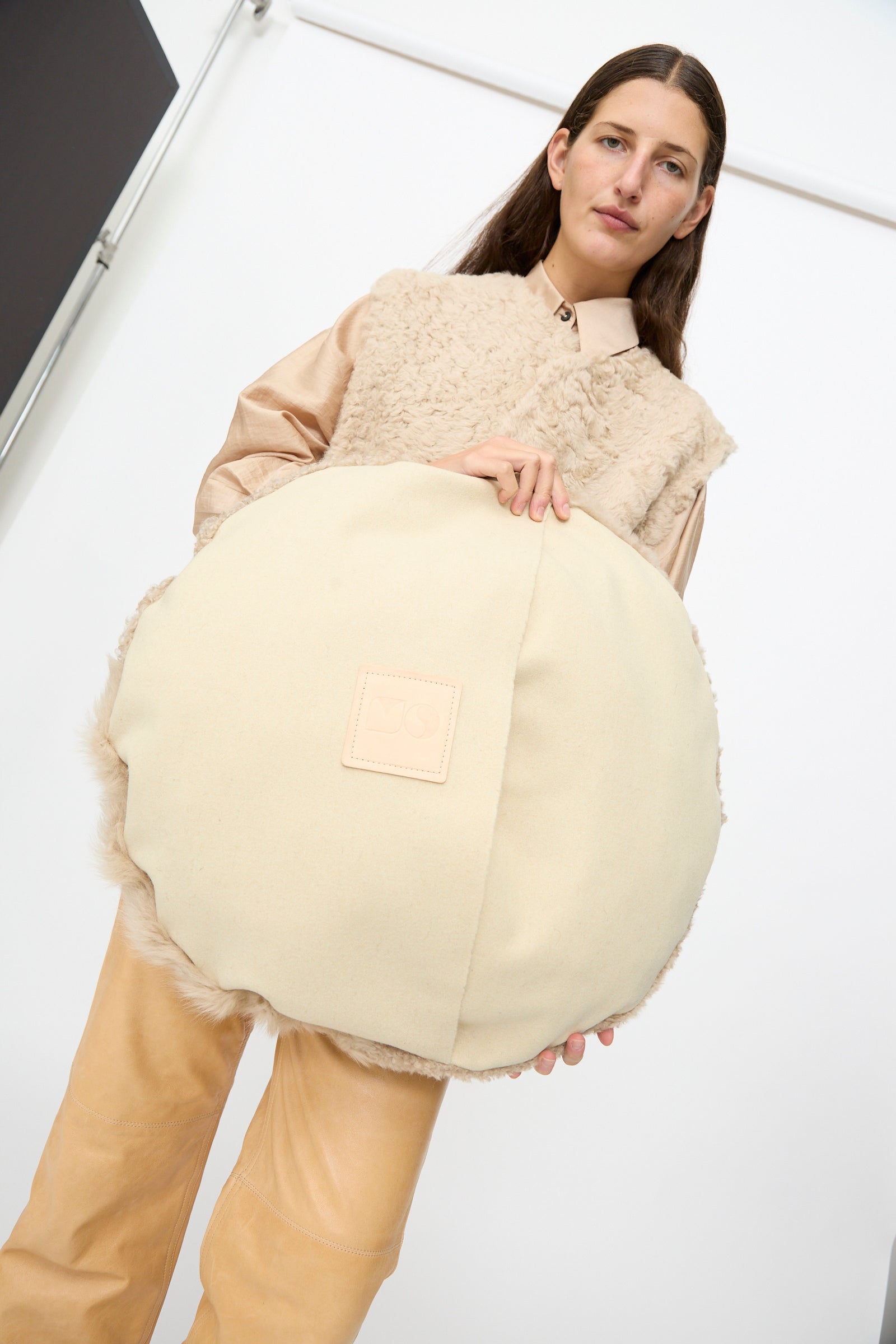 A person dressed in neutral tones holds the Milena Silvano Sheepskin Large Floor Pillow in Oat, featuring a stitched square patch, within a minimalist indoor setting.