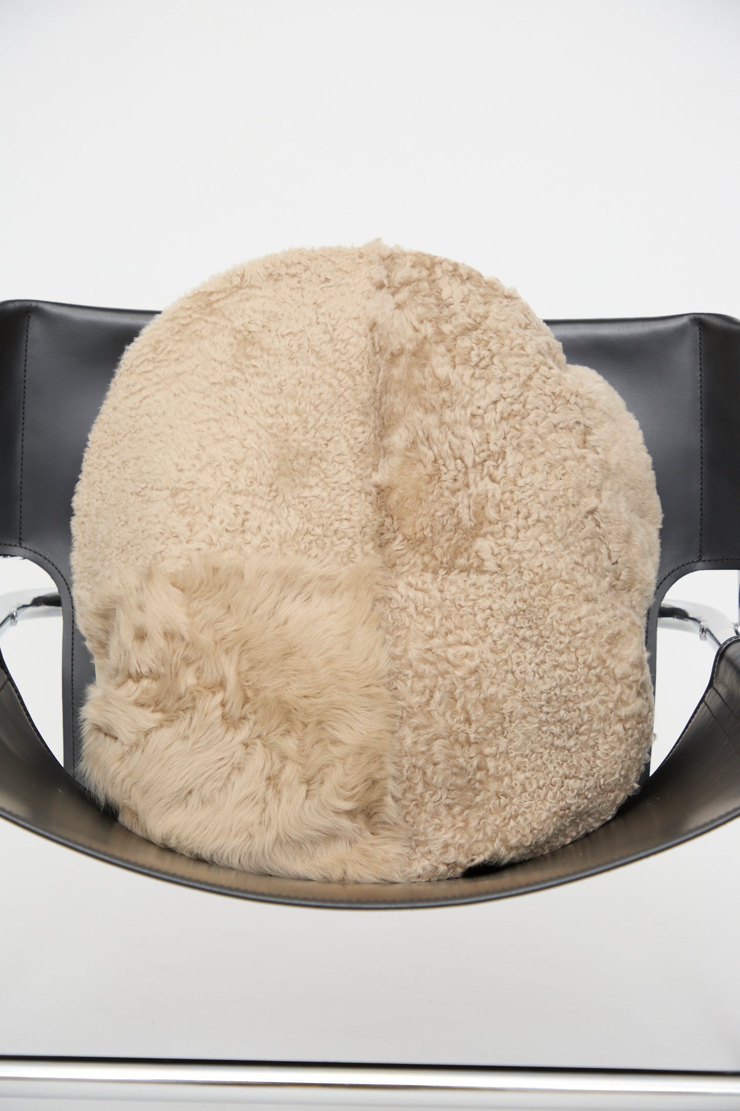 The Milena Silvano Sheepskin Large Floor Pillow in Oat, featuring round, two-toned beige and tan patchwork, is placed on a black leather chair with metal armrests against a plain white background.
