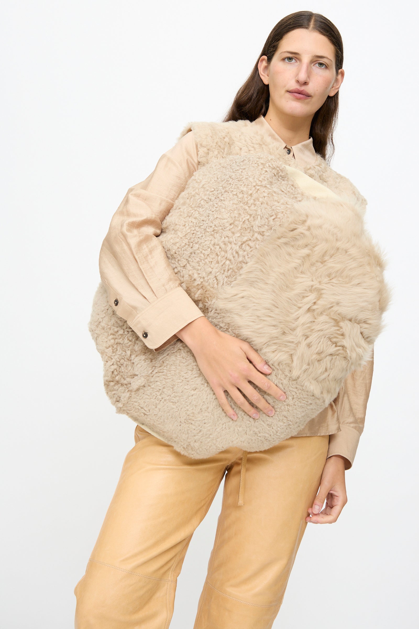 A person in tan clothing holds the Milena Silvano Sheepskin Large Floor Pillow in Oat in front of a plain white background.