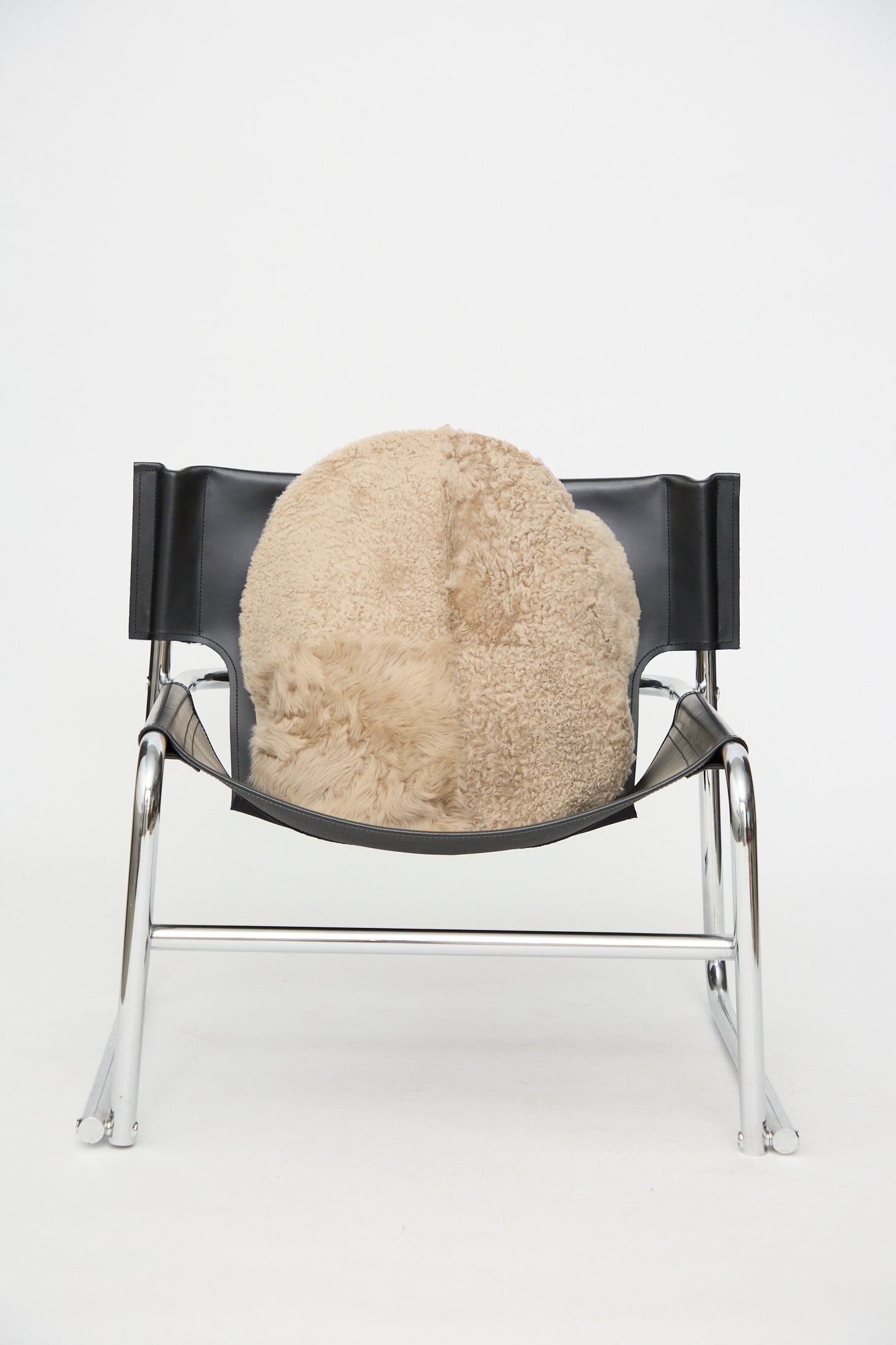 A Milena Silvano Sheepskin Large Floor Pillow in Oat, reminiscent of a patchwork sheepskin pillow, rests on the seat of a black leather and chrome chair that is centered on a white background.