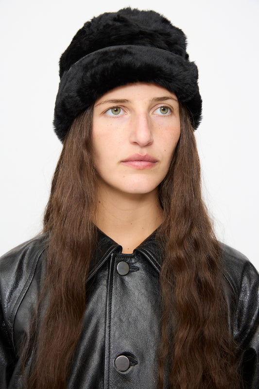 A person with long brown hair wears the Milena Silvano Sheepskin Pixie Hat in Black and a black leather jacket, looking slightly to the side against a plain background.