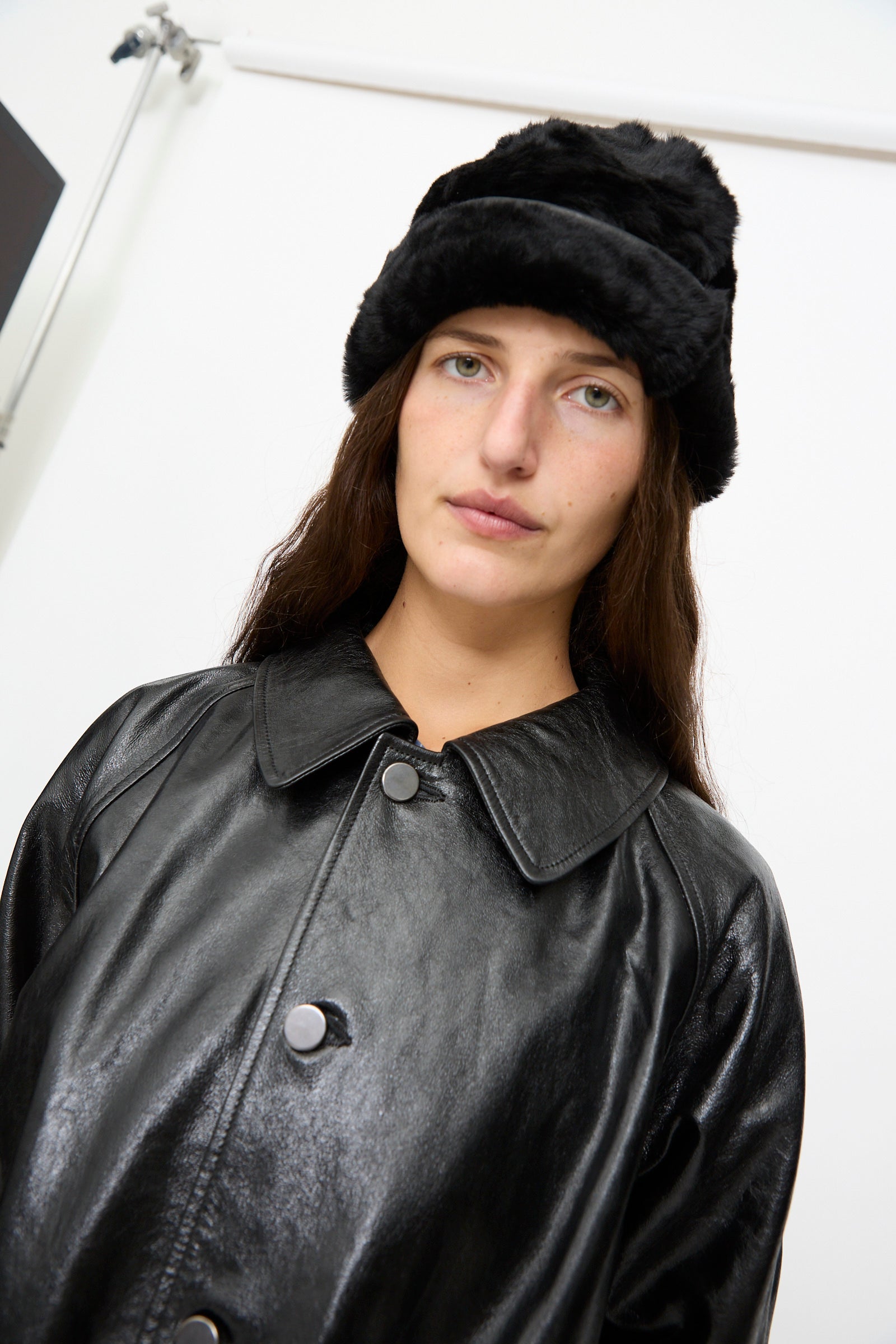 A woman wearing the Milena Silvano Sheepskin Pixie Hat in Black and a black leather coat stands against a plain white background, looking at the camera.