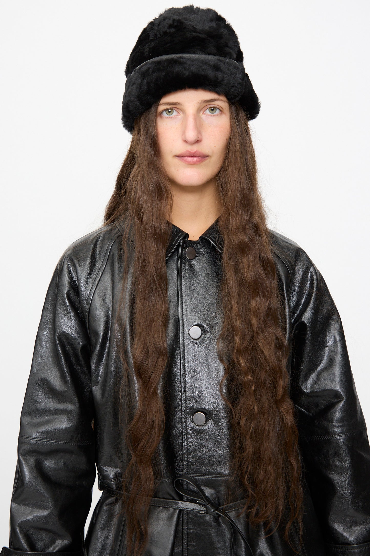A person with long brown hair wears the Milena Silvano Sheepskin Pixie Hat in Black and a black leather coat, standing against a plain white background.