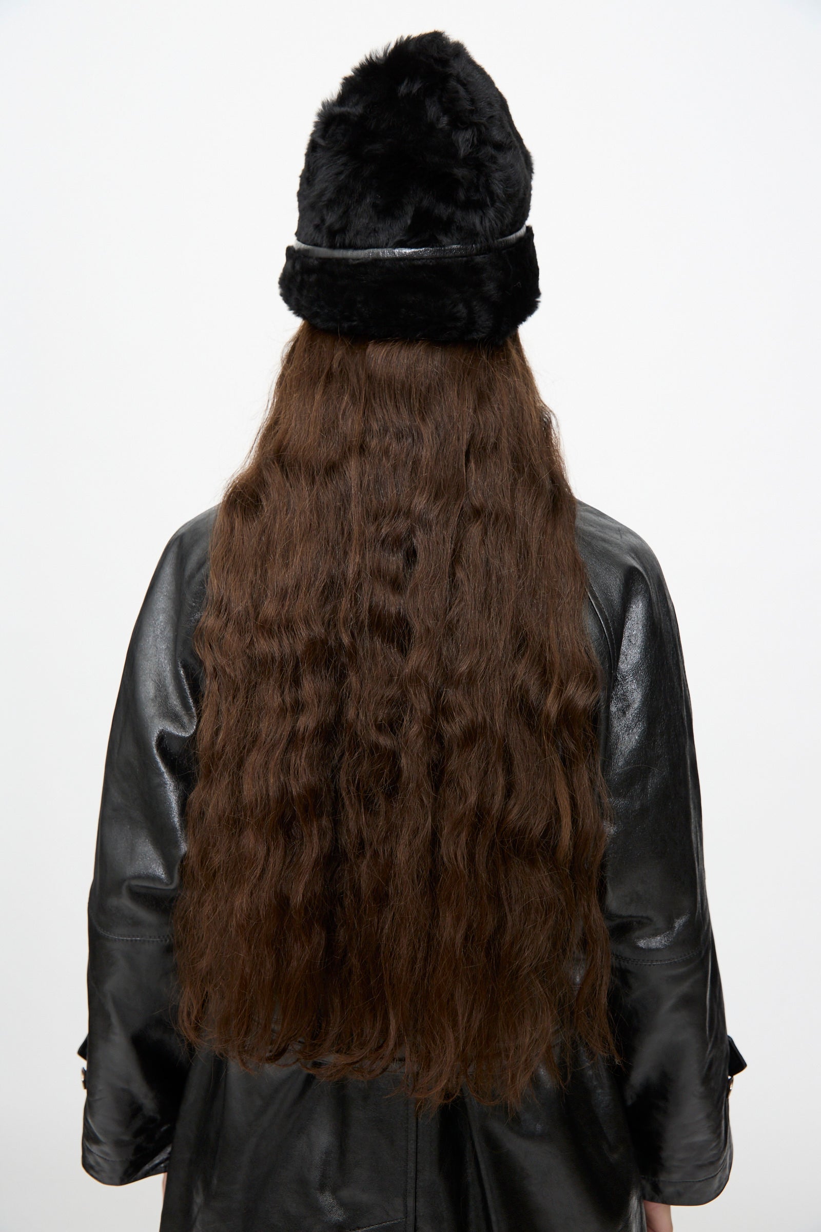 A person with long, wavy brown hair is shown from the back against a plain white background, wearing the Milena Silvano Sheepskin Pixie Hat in Black and a black leather coat.