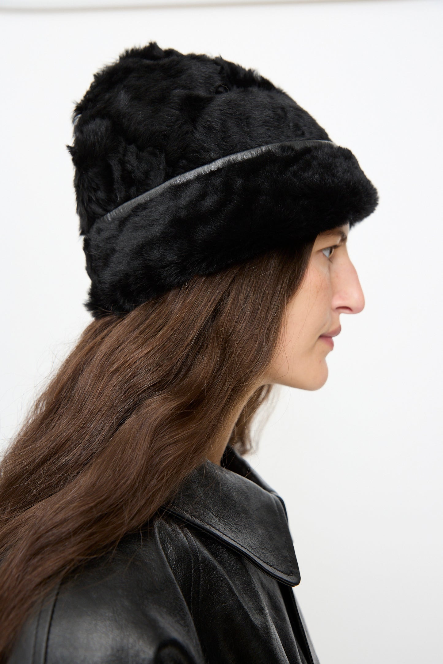 A person with long brown hair, shown in profile against a plain white background, wears the Milena Silvano Sheepskin Pixie Hat in Black and a black leather jacket.