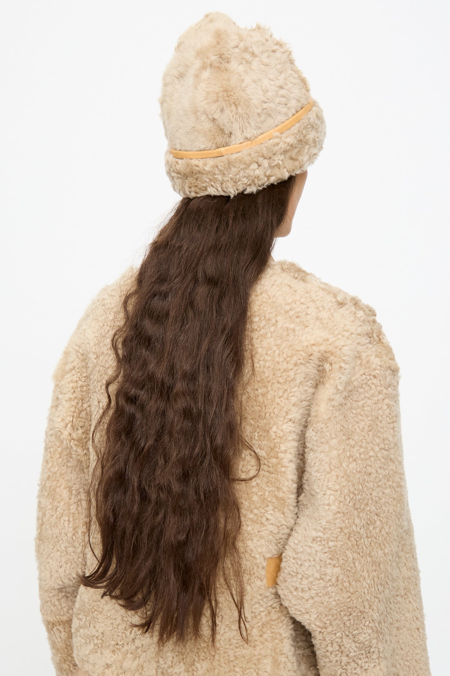 A person with long, wavy brown hair is seen from the back against a plain white background, wearing a beige faux shearling coat and the Milena Silvano Sheepskin Pixie Hat in Oat.