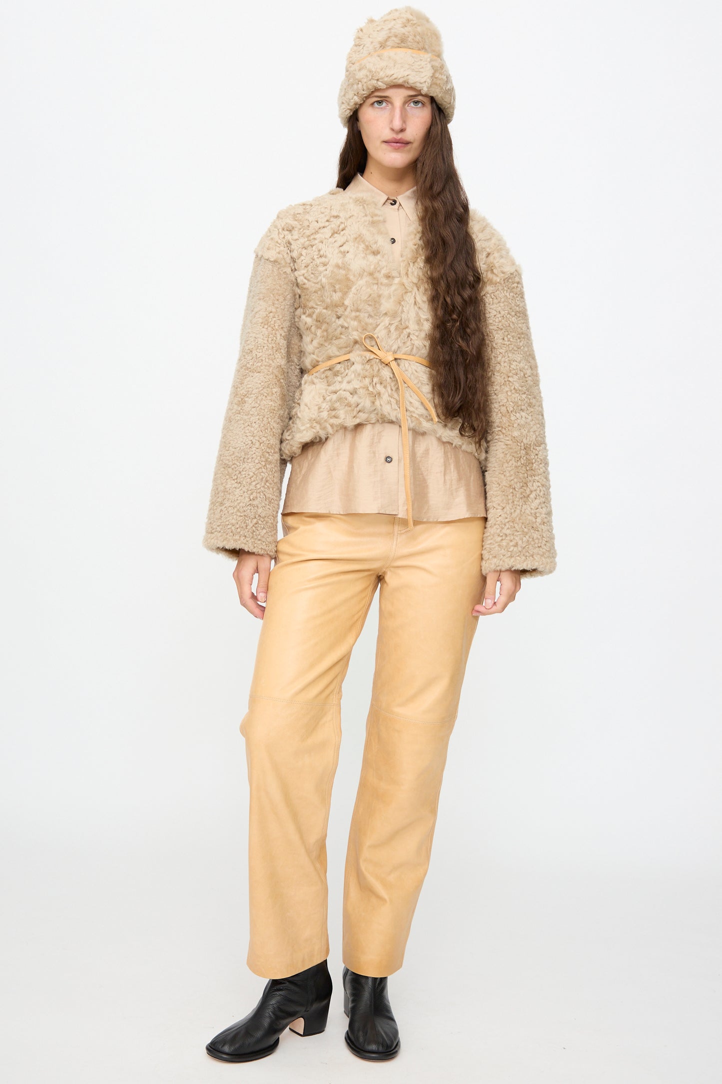 A person wearing the Milena Silvano Sheepskin Pixie Hat in Oat, a textured beige jacket, tan shirt, tan pants, and black ankle boots stands against a plain white background.