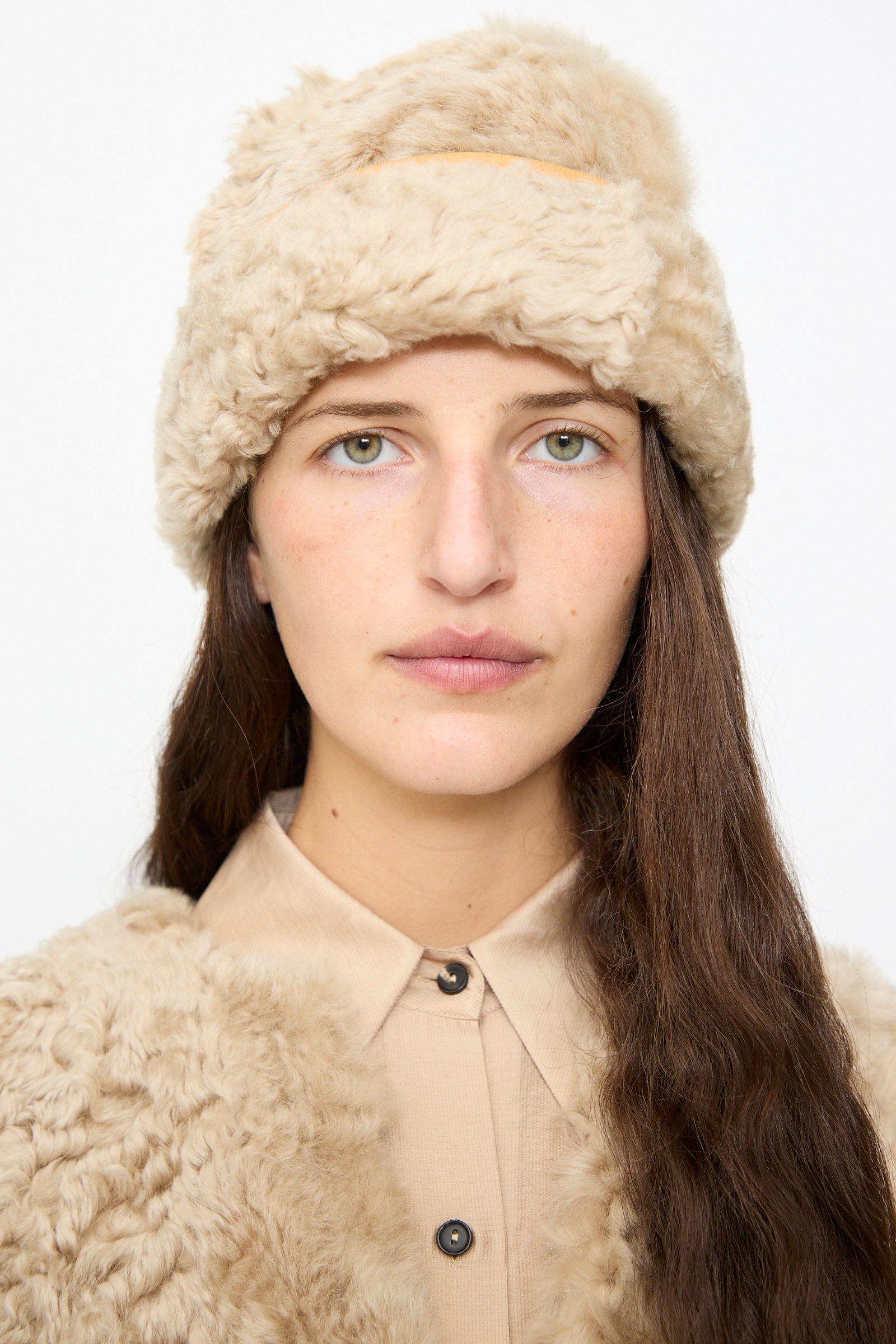 Wearing the Milena Silvano Sheepskin Pixie Hat in Oat, a person with long brown hair stands against a plain white background, pairing the stylish leather-trimmed fur hat with a tan faux fur coat and a beige button-up shirt.
