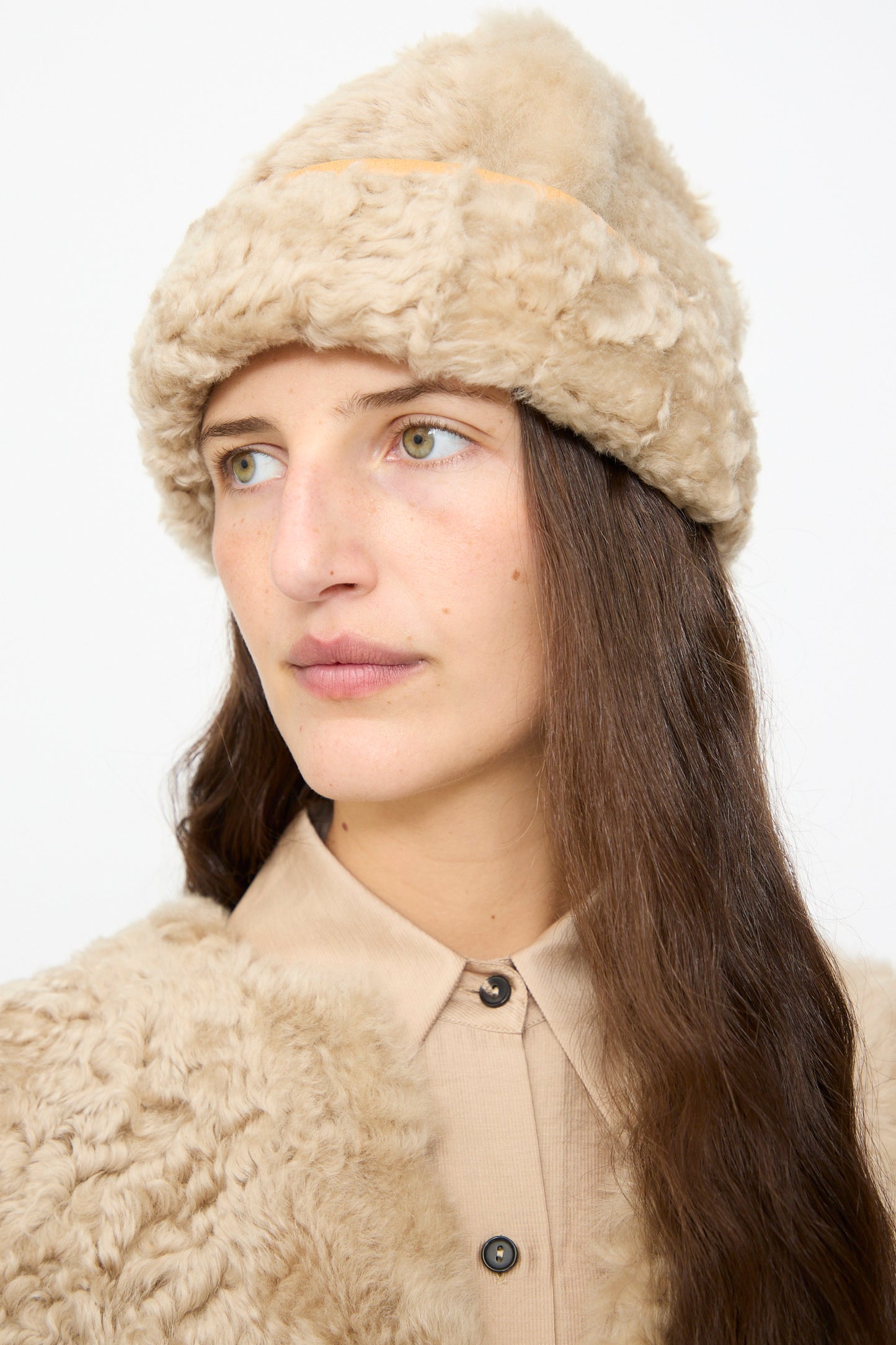 A woman wearing the Milena Silvano Sheepskin Pixie Hat in Oat and a matching beige coat over a beige button-up shirt, looking slightly to the left against a plain white background.