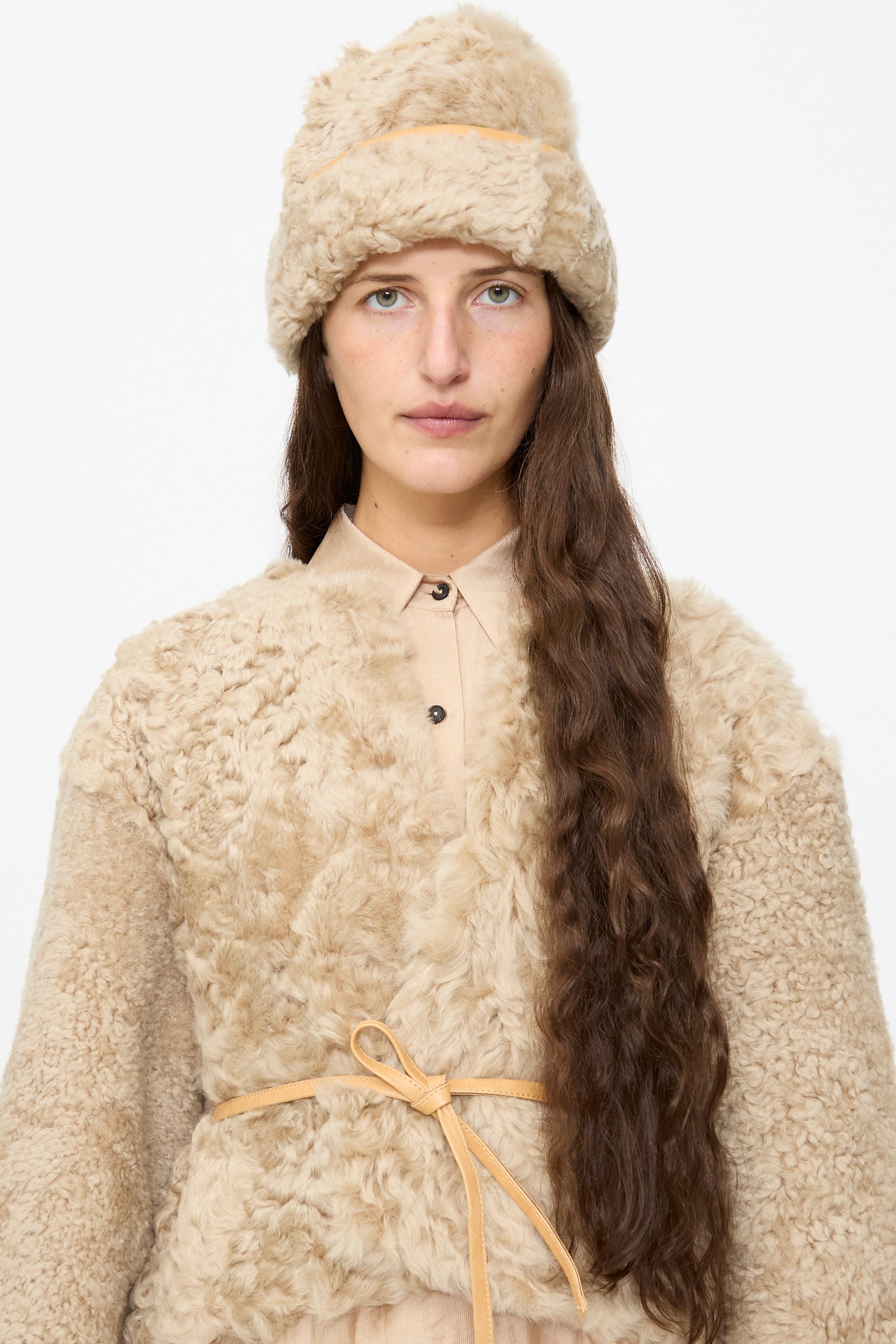 A woman models the Milena Silvano Sheepskin Pixie Hat in Oat, featuring a beige sheepskin design with leather trim, paired with a coordinating jacket as her long brown hair cascades down against a plain white background.