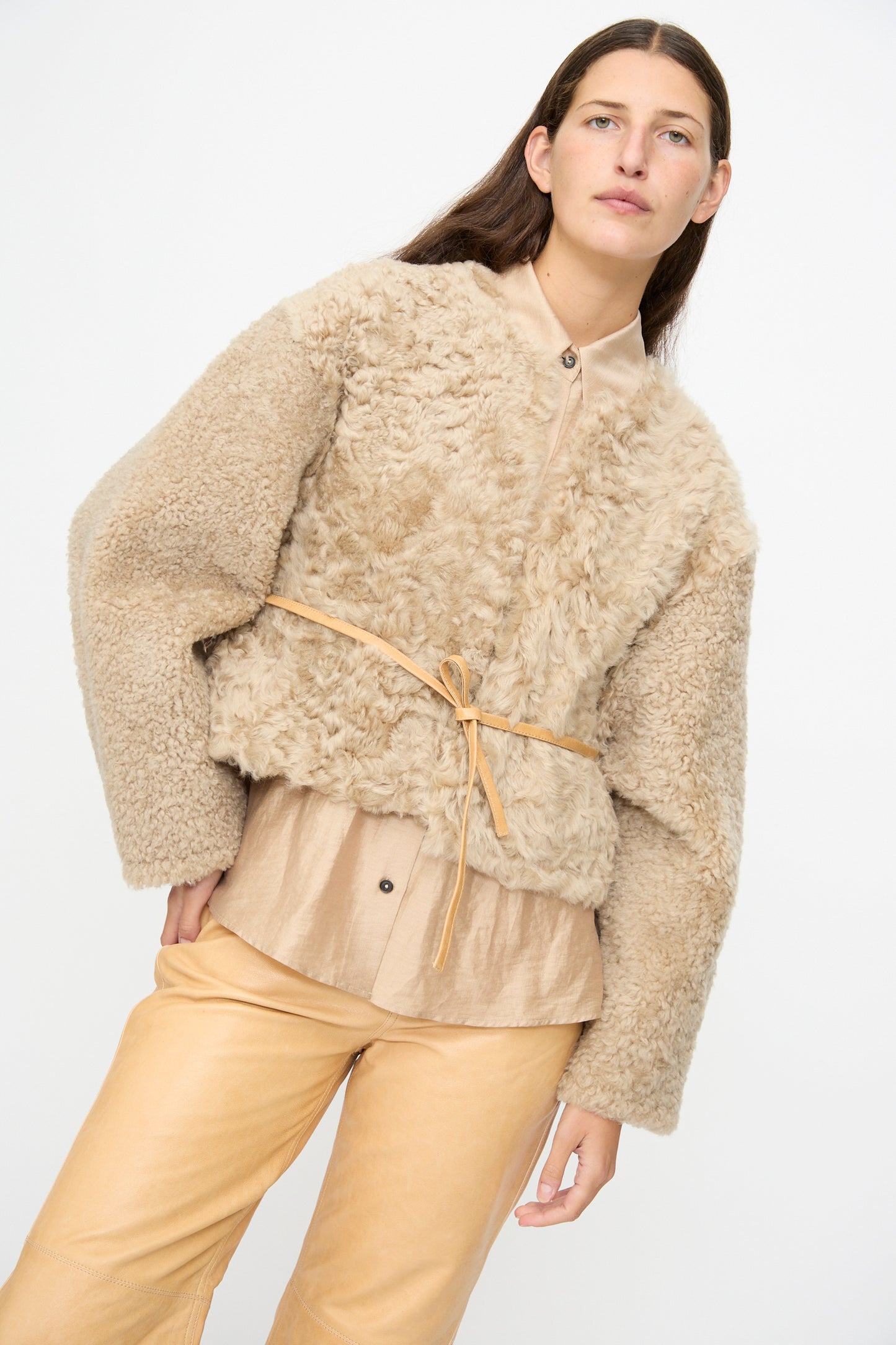 A person wearing the Milena Silvano Sheepskin Rainer Jacket in Oat, paired with a light tan button-up shirt and matching tan pants, stands against a plain white background.
