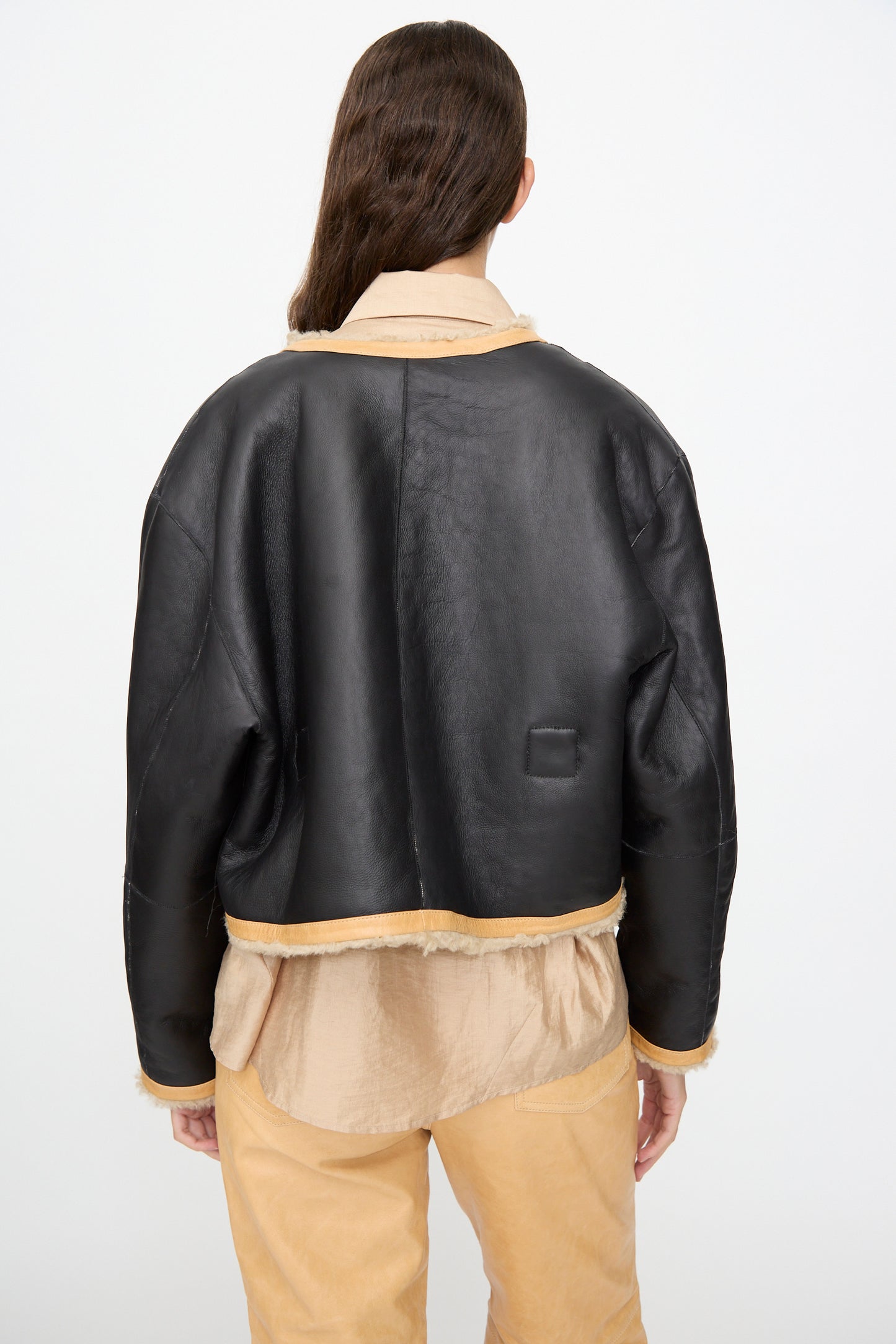 A person with long brown hair is seen from behind against a plain white background, wearing the Milena Silvano Sheepskin Rainer Jacket in Oat over a beige shirt and pants.