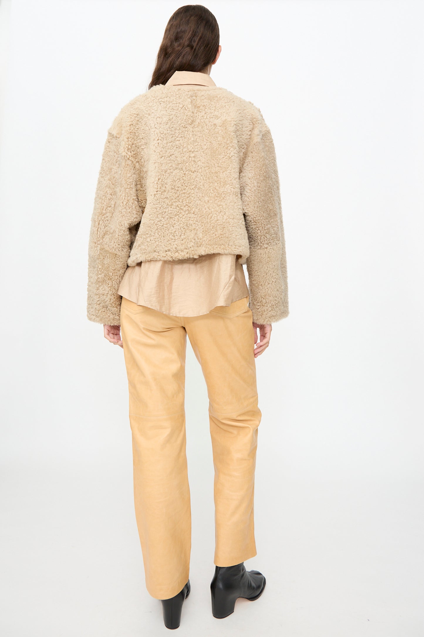 A person stands facing away, wearing the Milena Silvano Sheepskin Rainer Jacket in Oat—with leather waist ties—paired with a tan shirt, light tan pants, and black ankle boots against a plain white background.