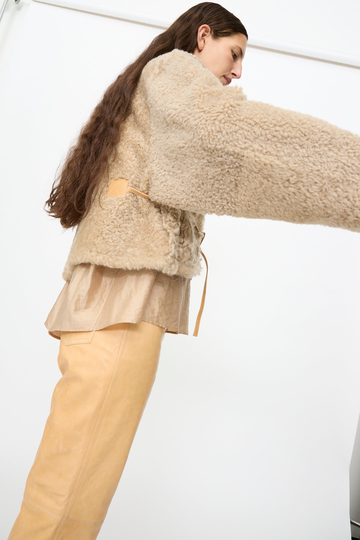 A woman with long brown hair wears the Milena Silvano Sheepskin Rainer Jacket in Oat, paired with a tan blouse and matching pants. She stands against a plain white background with her arms slightly extended.