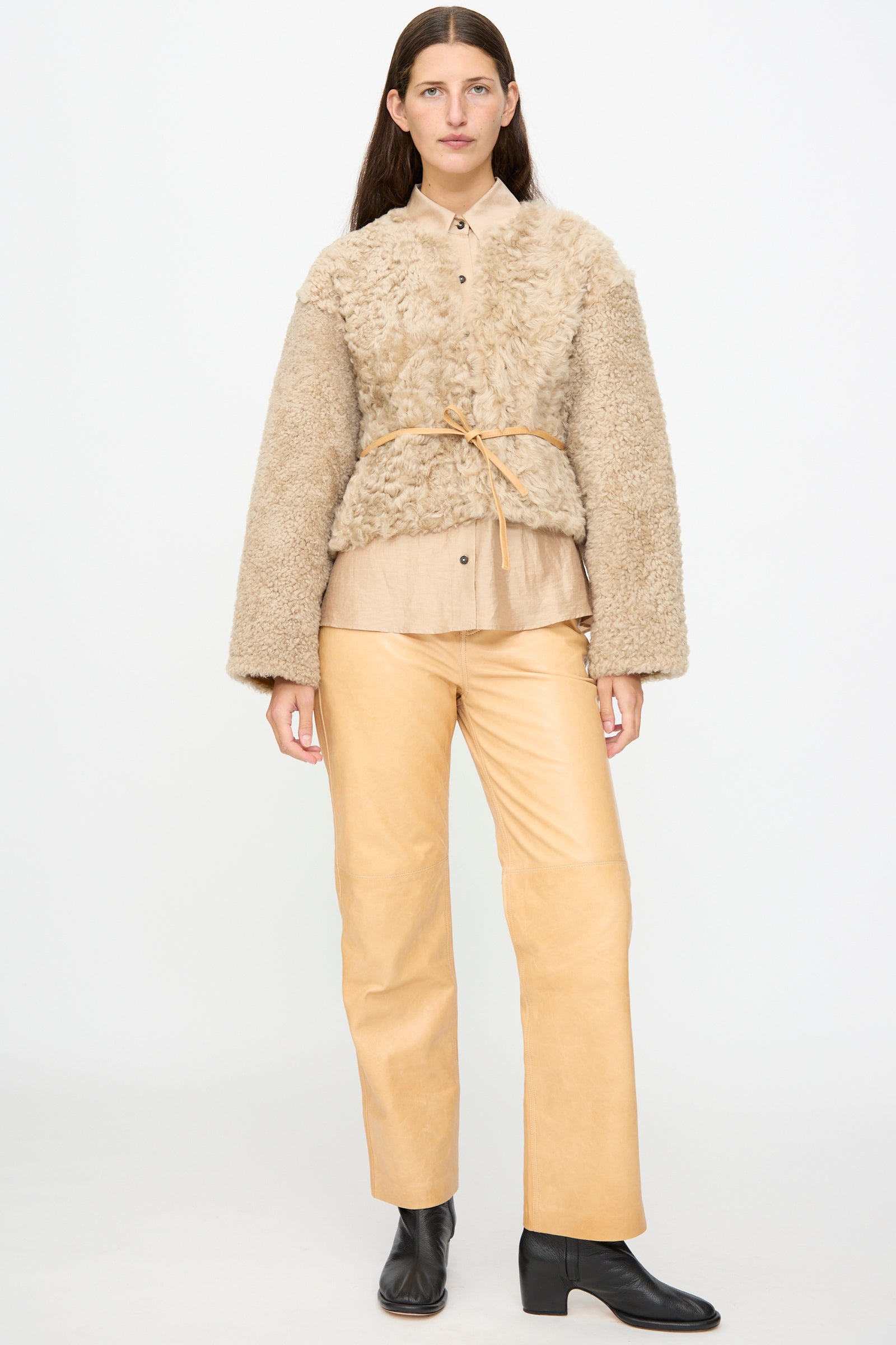 A woman stands against a plain background, wearing the Milena Silvano Sheepskin Rainer Jacket in Oat with leather waist ties over a tan shirt, paired with light tan pants and black ankle boots.
