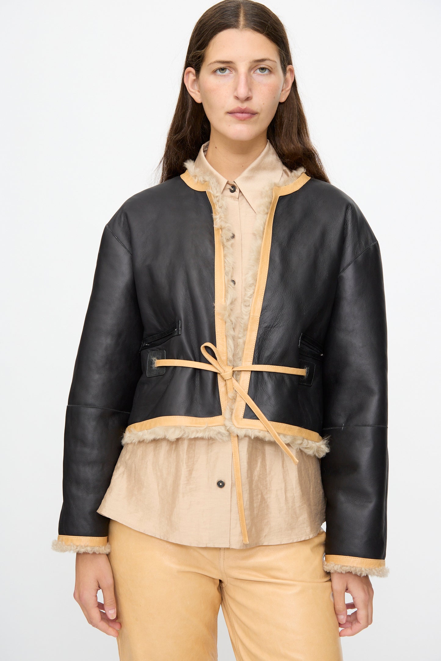 A woman wearing the Milena Silvano Sheepskin Rainer Jacket in Oat, paired with a beige button-up shirt and light tan pants, stands against a plain light background.