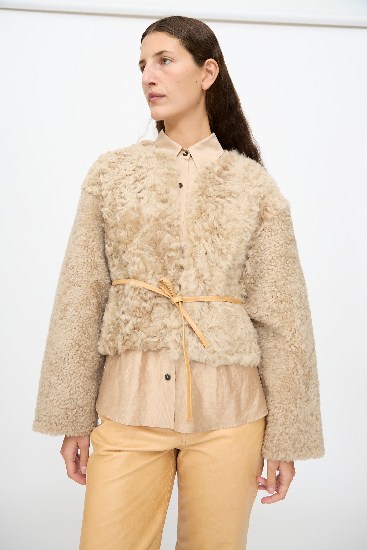 A woman wearing the Milena Silvano Sheepskin Rainer Jacket in Oat, styled over a button-up shirt and light tan pants, stands against a plain white background.
