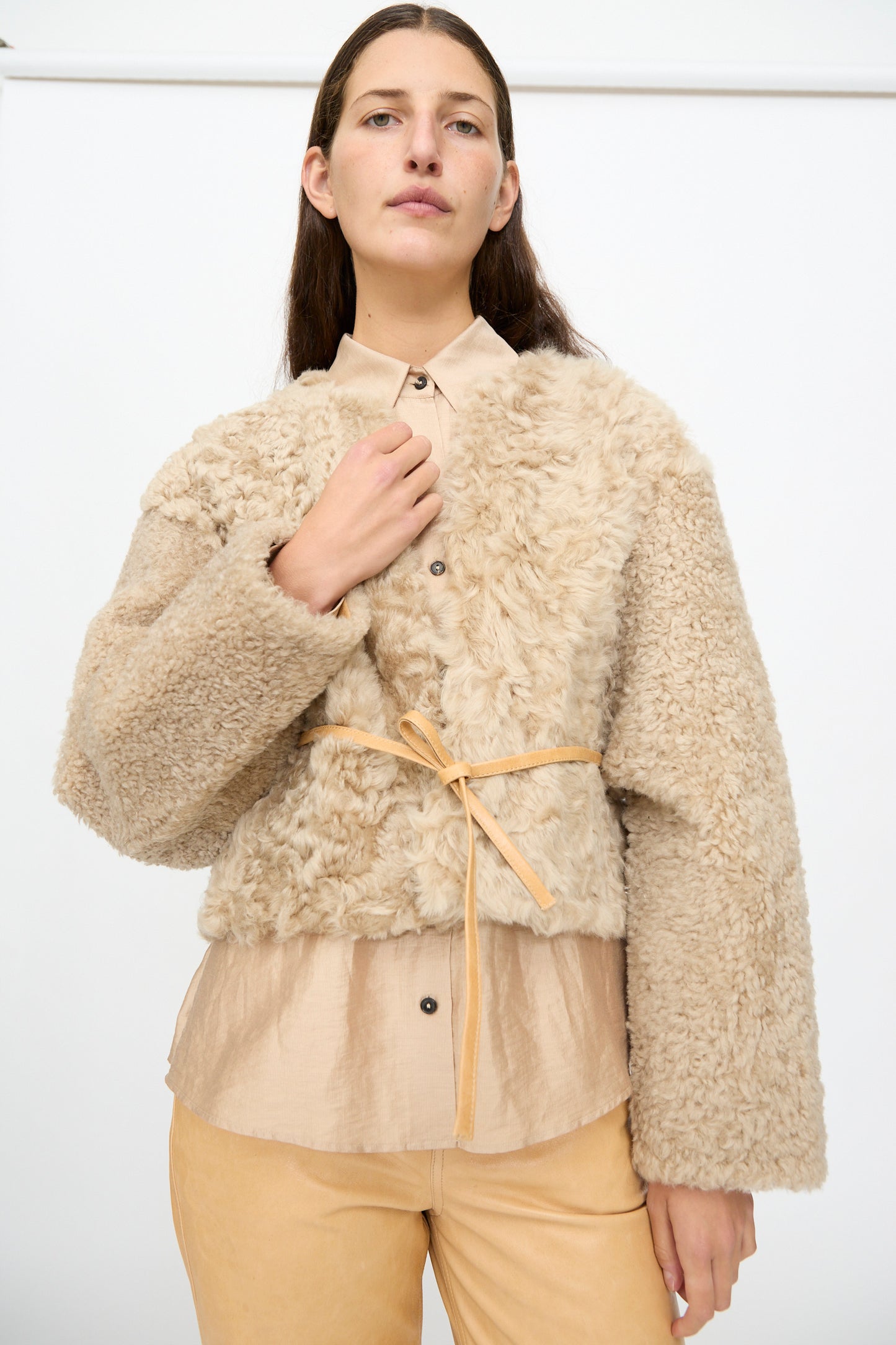 A person with long brown hair wears the Milena Silvano Sheepskin Rainer Jacket in Oat, tied at the waist over a tan shirt and tan pants, standing against a plain white background.