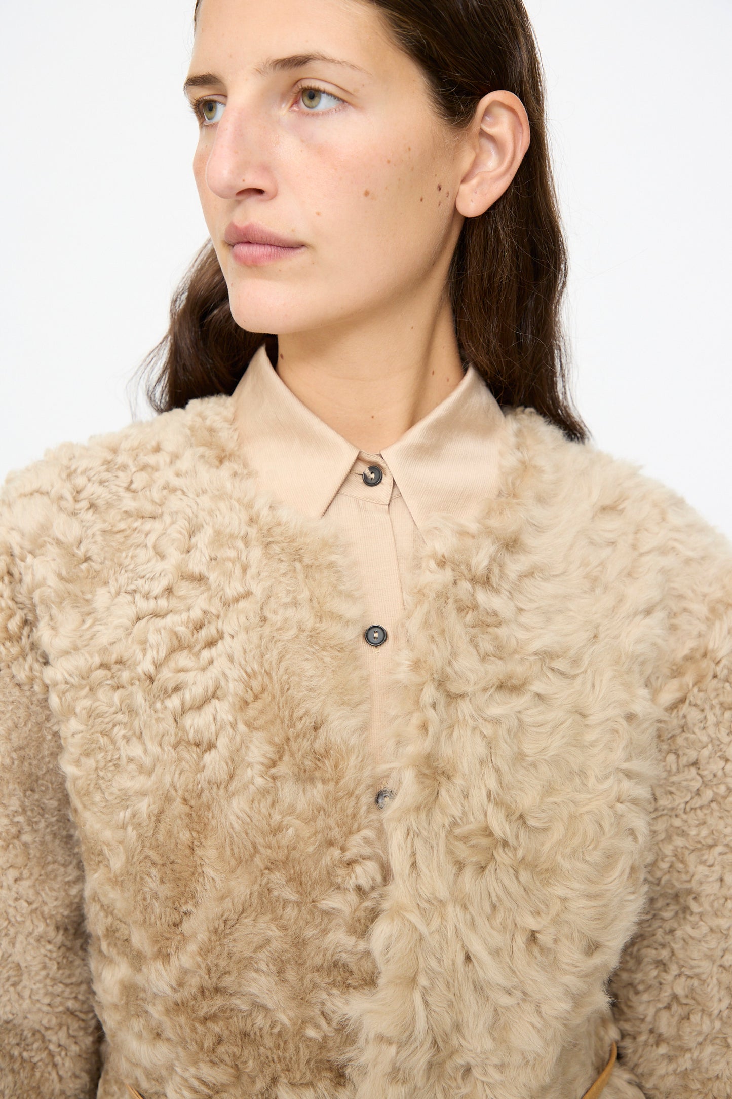 A woman with long brown hair models the Milena Silvano Sheepskin Rainer Jacket in Oat—a textured, light tan cropped sheepskin piece featuring chic leather waist ties—worn over a beige collared shirt, as she gazes slightly to the side against a plain background.