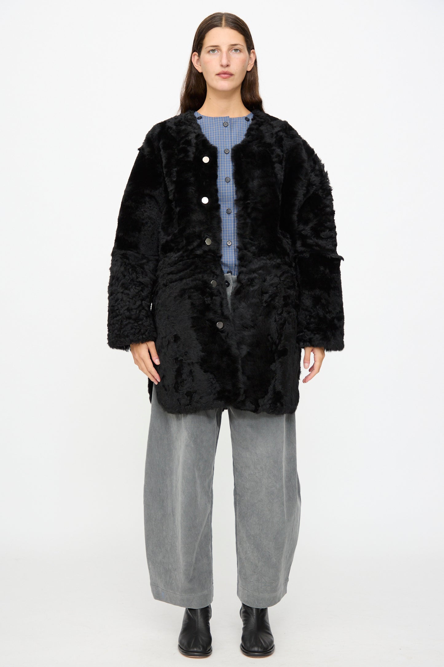 A person stands facing forward, wearing the Milena Silvano Sheepskin Reversible Liner in Black over a blue shirt and gray wide-leg pants with black boots against a plain white background.